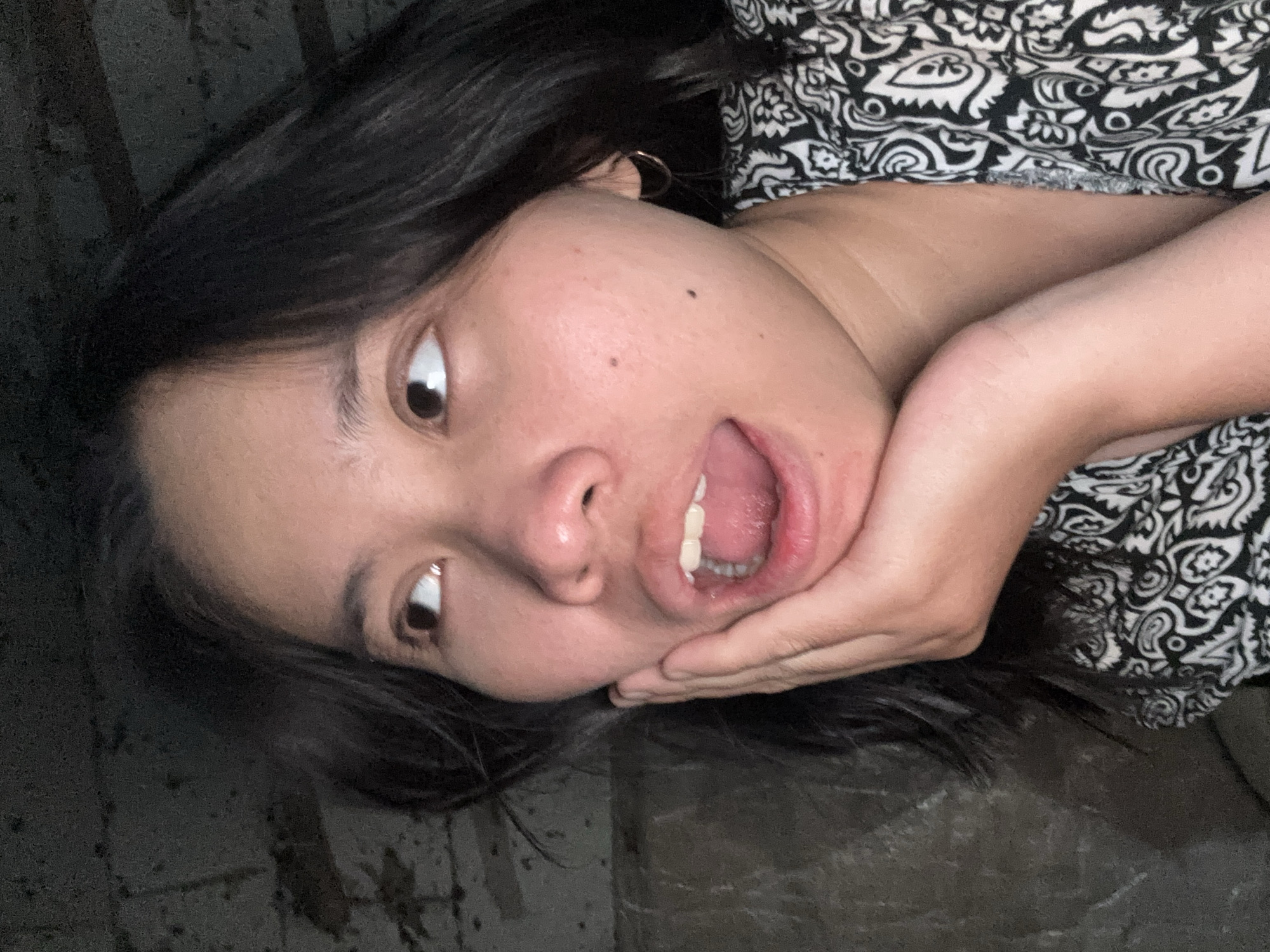 A young Asian woman with dark hair and a patterned shirt is looking up and to her left, with her mouth open in surprise and her hand on her cheek. She appears to be in an indoor setting with a rough, dark wall behind her. The lighting is dim, and the focus is on her face. The location is not discernible from the image.