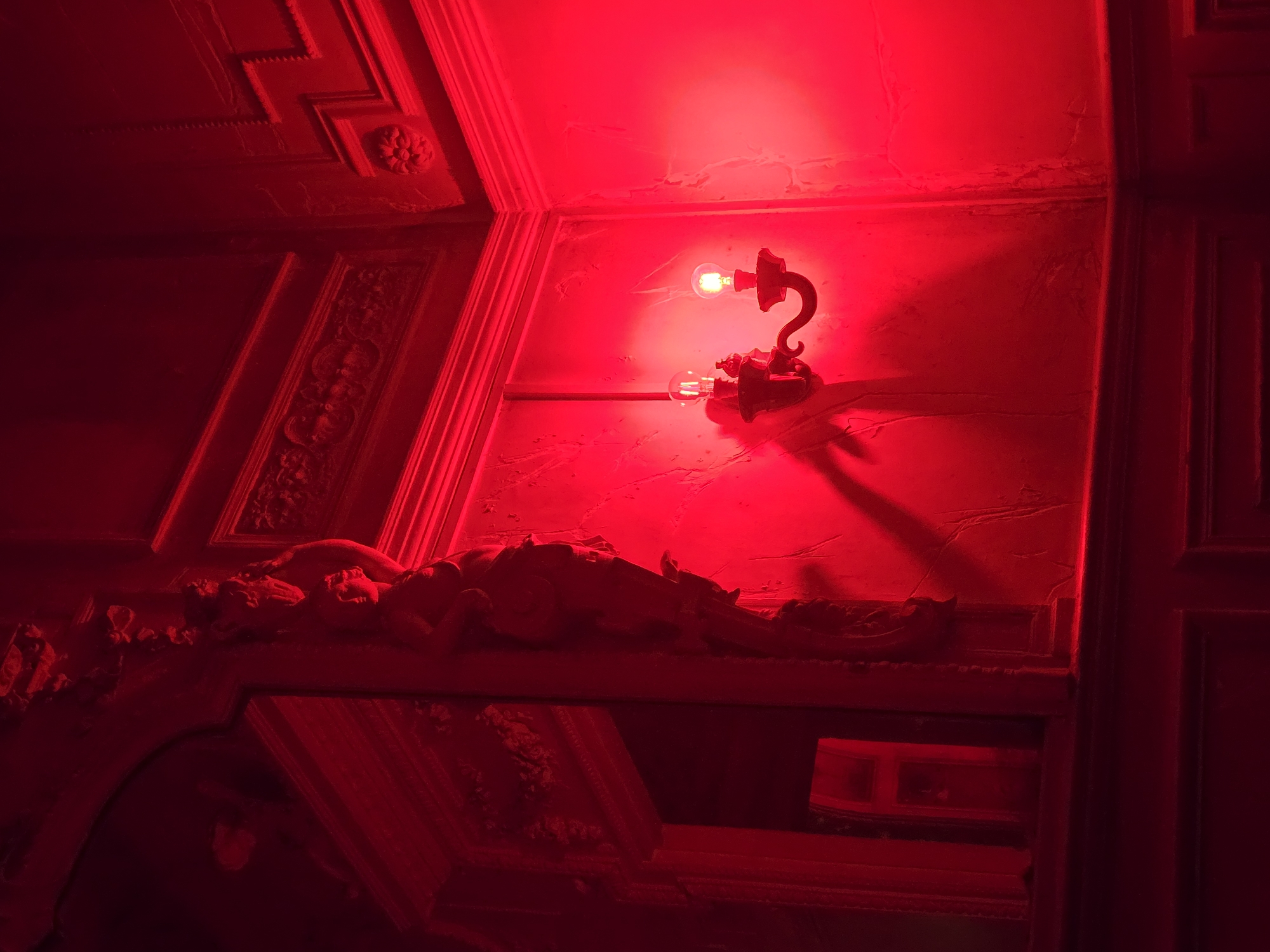 A dimly lit interior features ornate architectural details bathed in a dominant red hue. A light fixture with an exposed bulb is mounted on a wall with cracked or textured plaster. Below, what appears to be a decorative mantelpiece displays a horizontal figure, possibly a sculpture or relief, that is draped or reclining. The surfaces suggest historical craftsmanship. No persons are visible. No location is known.