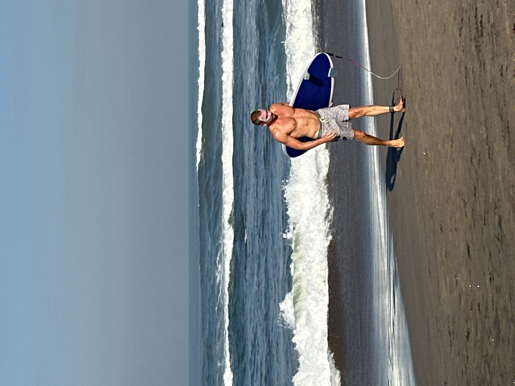 A shirtless male, appearing to be in his 30s with a muscular build, walks barefoot on a dark sandy beach in North Kuta, Indonesia, carrying a blue boogie board or small surfboard under his right arm. He wears patterned grey swim shorts and has white sun protection cream visibly applied to his forehead, nose, and cheeks. A leash, attached to the board, is secured around his left ankle. The man faces slightly towards the left of the frame, with his gaze directed forward. In the background, the vast blue-grey ocean features multiple lines of white-capped waves rolling towards the shore. The sky above is a clear, hazy blue, transitioning from lighter blue at the horizon to a deeper blue overhead. The wet sand near the waterline reflects the sky, while the sand further up the beach is drier and exhibits subtle variations in texture and color.