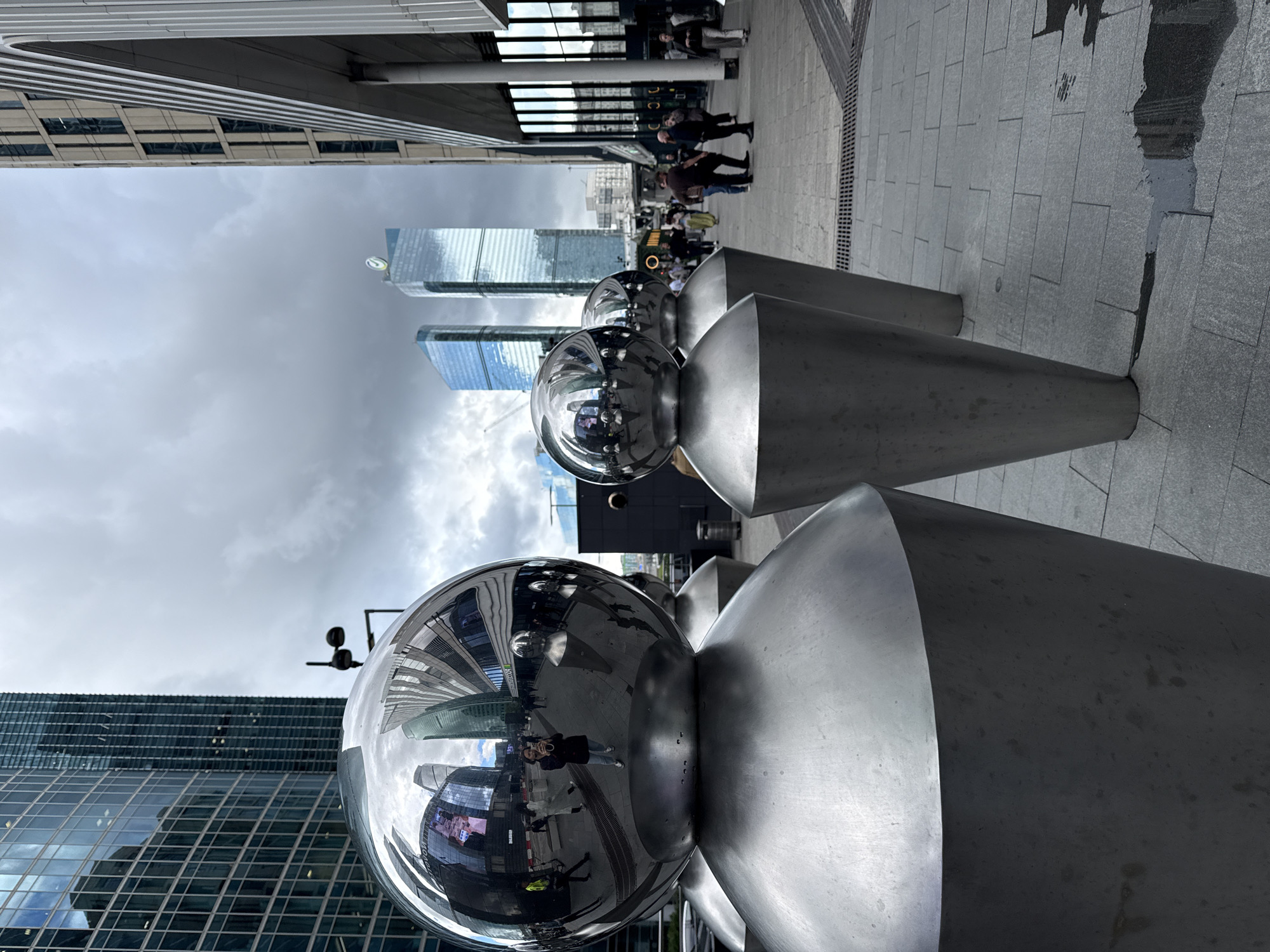 The image shows a street scene in Moscow City, Russia, featuring a row of polished metal sculptures that resemble inverted cones topped with spheres. These sculptures are positioned on a paved walkway. The sphere on the leftmost sculpture prominently reflects the surrounding environment, including tall glass-fronted skyscrapers, a cloudy sky, and several people walking in the distance. Among the reflected figures, a woman is visible taking a photograph of herself. Other sculptures are visible further down the walkway, receding into the distance. In the background, modern high-rise buildings, characteristic of a financial district, dominate the skyline. People are seen walking and interacting on the pedestrianized area. The lighting suggests an overcast day.