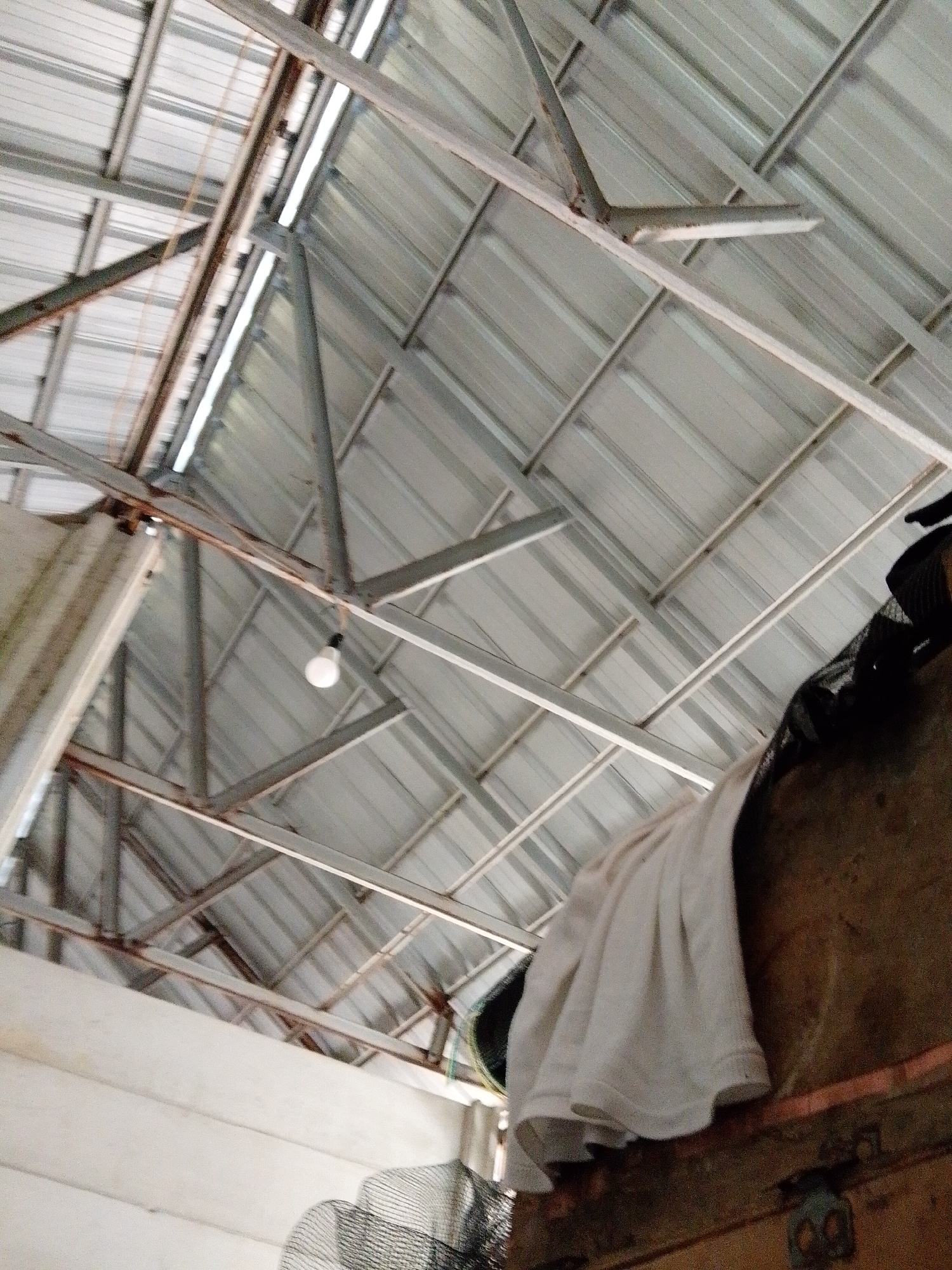 The image depicts the interior of a structure in Bukit Ketok, Indonesia, likely a storage area or shed, characterized by a metal roof supported by a framework of steel beams. A single lightbulb hangs from the structure. Visible in the foreground are various objects, including a trunk or wooden storage box draped with a white cloth. A mesh-like object is also present.