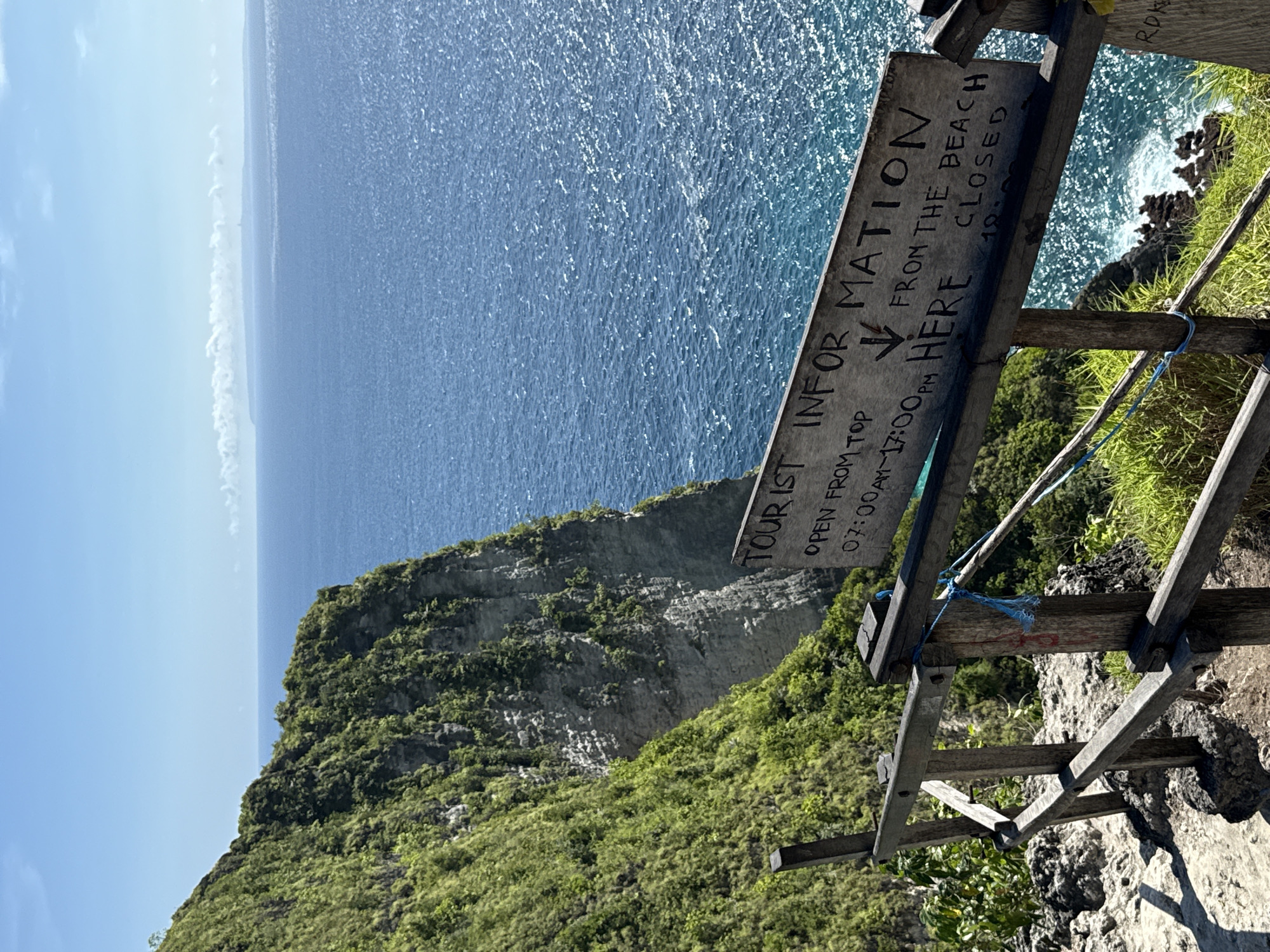 Captured at Bunga Mekar, Indonesia, the image presents an elevated perspective of a cliffside meeting the ocean. Lush vegetation blankets the cliff, transitioning into rocky terrain at its edge. The ocean stretches towards the horizon, displaying varying shades of blue. A weathered wooden sign with the inscription "TOURIST INFORMATION" stands prominently, providing information such as "OPEN FROM TOP 07:00AM~17:00PM" and "FROM THE BEACH CLOSED." The sign is supported by a basic wooden structure with a blue rope detail, set against the backdrop of the cliff and the ocean.