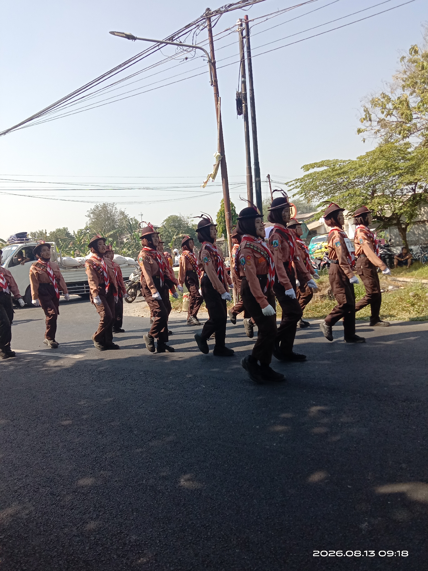 In the city of Sukoiber, Indonesia, a group of uniformed scouts are marching in a procession on a paved road. The scouts, predominantly young individuals, are wearing brown uniforms with red and white neckerchiefs, and brown hats adorned with feathers. They are marching in formation, with some holding flags or banners, although these are not clearly visible. A white truck with a covered cargo area is visible to the left of the procession. The environment appears to be a suburban or rural area, with trees and foliage lining the right side of the road, and utility poles and wires overhead. The sky is clear and bright, indicating daytime. The overall scene suggests a ceremonial event or a parade.