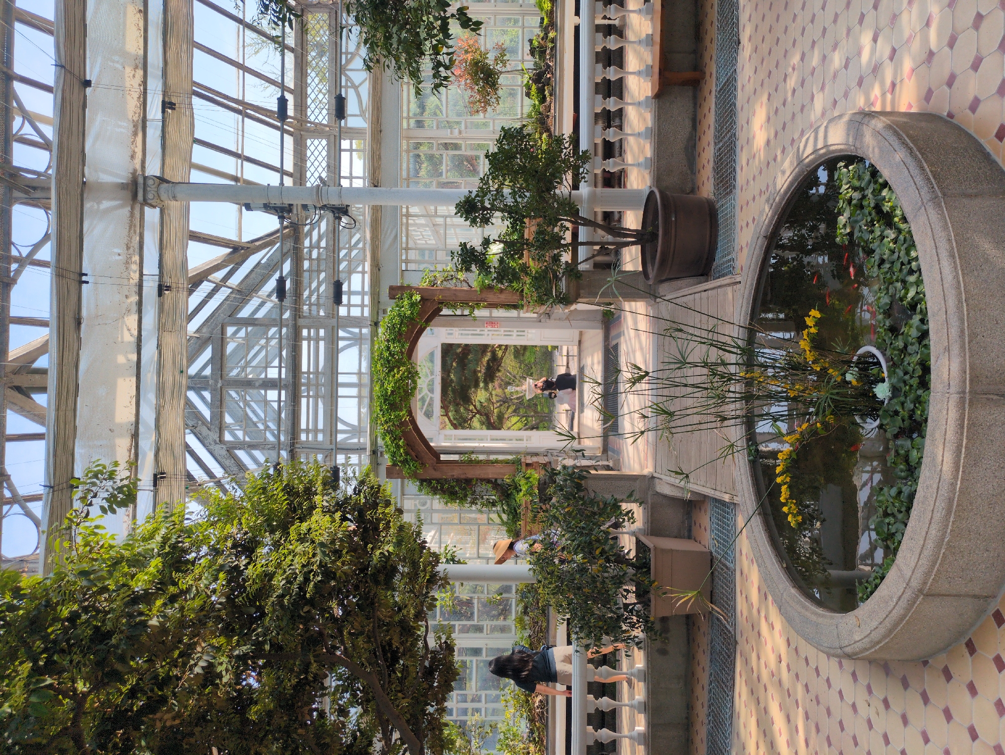 Inside a greenhouse in Seoul, South Korea, a large circular fountain dominates the foreground with water, greenery, and visible goldfish. The walkway leads toward an arched entrance adorned with climbing plants. Two people are visible in the distance, appearing to be a couple. One woman with a hat stands near a white railing in the middle ground. The greenhouse structure is made of white beams and glass panels, allowing bright sunlight to enter. The flooring is tiled in a patterned design. Numerous potted plants and trees add lushness to the interior space.