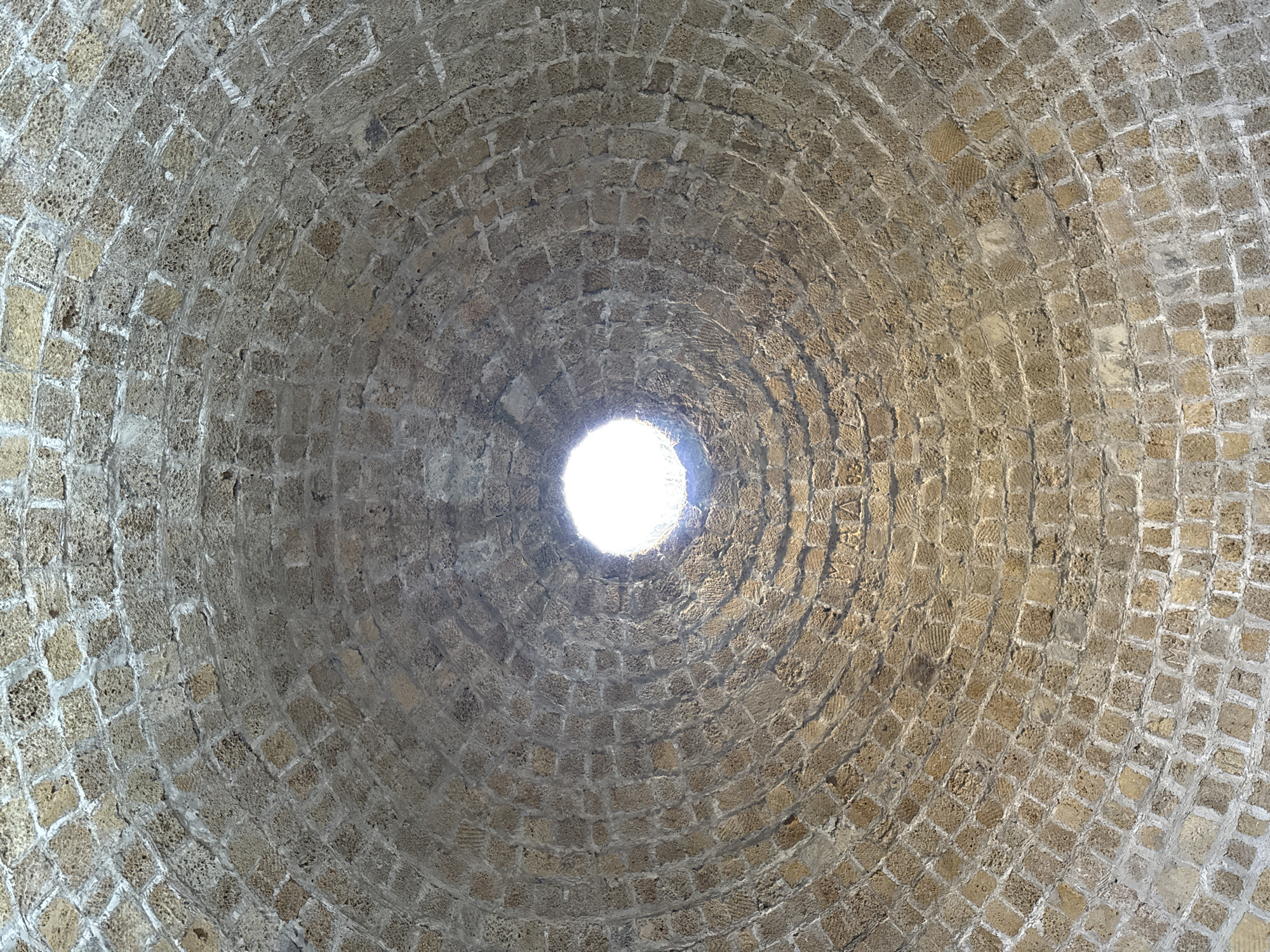 This media file captures an upward view of a domed ceiling constructed from rough-hewn, small, square-shaped stones. The stones are arranged in concentric circles, creating a spiral pattern that converges towards a circular opening at the apex of the dome. This opening allows a bright light to enter, illuminating the interior and casting subtle shadows on the textured stone surfaces.
The material of the dome appears to be porous and weathered, suggesting age and exposure to the elements. The overall impression is of an ancient structure, possibly a well, cistern, or part of a historic building. The rough texture of the stones and the irregular mortar lines contribute to a rustic and somewhat primitive aesthetic.
There are no people or overt objects visible in the frame, and the focus is entirely on the architectural detail of the dome. The lighting suggests it is daytime, with the bright light from the opening indicating clear or at least bright conditions outside. The location context of Derbent, Russia, hints at a historical or cultural site within that region.
On the stone surface, faint, carved markings are visible in some areas, particularly towards the right side of the image. These appear to be inscriptions or graffiti, though their legibility is limited due to the texture and lighting. One visible inscription, near the top right, contains the letters "VA" and "93" suggesting a date or initials. These markings add another layer to the historical narrative of the structure. The emotion evoked by the image is one of awe and perhaps curiosity about the history and purpose of this ancient dome.