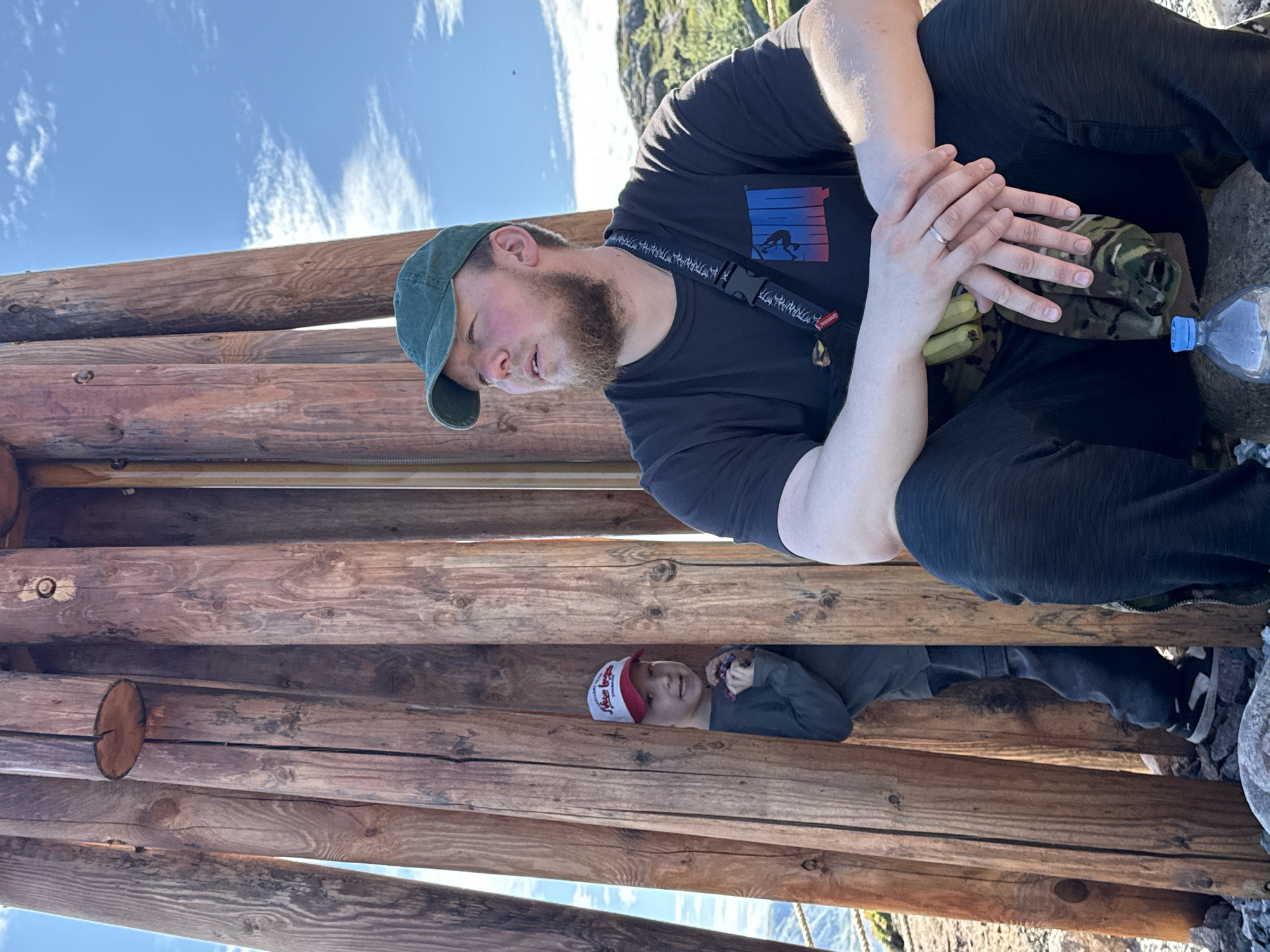 In Teriberka, Russia, a young boy with a red and white baseball cap is partially hidden behind a wooden structure. He is looking towards the camera with a slight smile. To his right, a man with a beard and a green baseball cap is sitting down, looking off to the side. He is wearing a black t-shirt with a graphic design and a crossbody strap. His hands are clasped in his lap, and he is wearing a ring. A bottle of water is placed in front of him on the ground. The background shows a clear blue sky with some clouds and a rocky, grassy landscape. The wooden structure appears to be made of large, vertical logs.