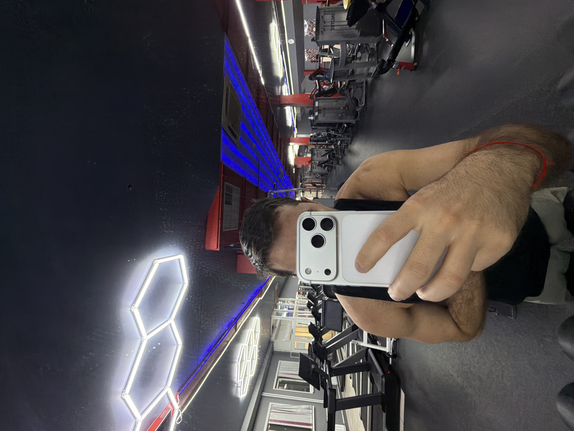 A man takes a selfie in a gym in Krasnoyarsk, Russia. He holds a white smartphone in front of his face, obscuring his features except for his hair and part of his forehead. He is wearing a black top and a red string is visible around his right wrist. The gym has a black ceiling and gray floor. Gym equipment is visible in the background, including weight machines and lighting fixtures. The walls feature geometric neon lights.