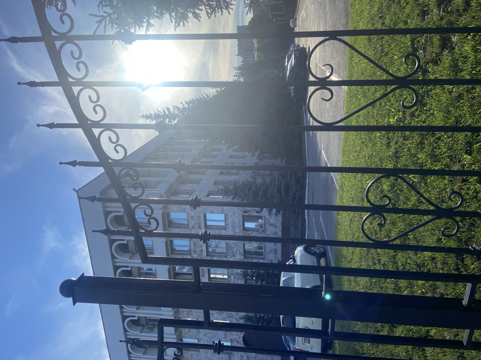 The image depicts a view through a black wrought-iron fence, adorned with heart-shaped embellishments, towards a modern administrative building with arched windows and light-colored stone facade. The building is set back from the fence, with a paved parking lot in between, where several cars are parked. Evergreen trees are visible along the side of the building and in the background. The scene is bathed in bright sunlight, with the sun appearing high in a clear blue sky with scattered clouds, causing lens flare in the image. The foreground shows a patch of green grass. The location is identified as the Republic of Bashkortostan National Library in Ufa, Russia.