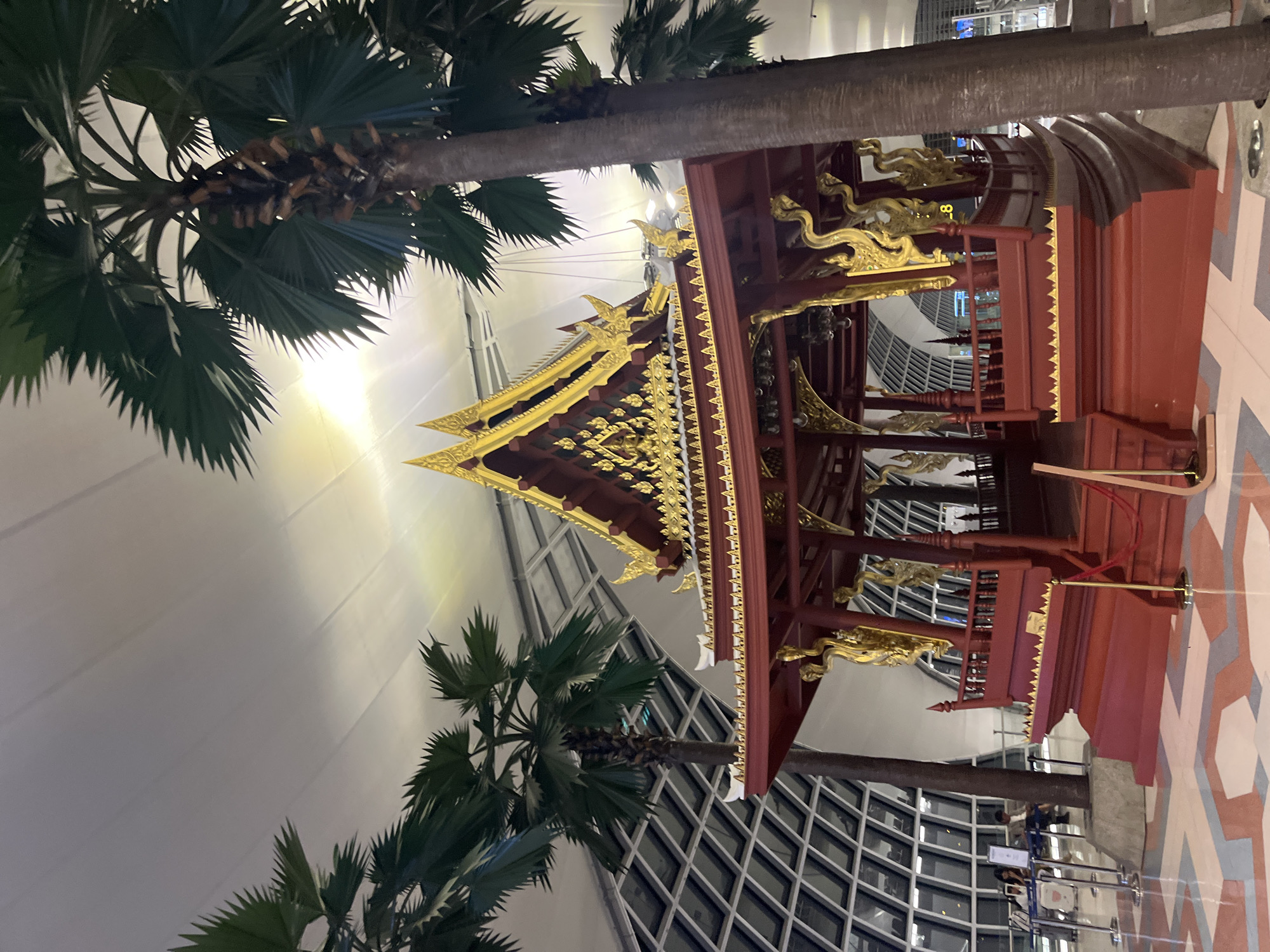 The image shows a traditional Thai pavilion structure, painted crimson red and adorned with gold accents, located indoors. The pavilion features an ornate, multi-tiered roof with intricate golden carvings and decorative finials. It has an open-air design with a railing around its upper level. Two palm trees with large fronds frame the pavilion, one on each side, with their trunks partially obscuring the structure and the background. The ceiling above is a large, white, curved surface with visible structural elements and lighting. In the background, a modern architectural element with a grid pattern of windows is visible. The floor is tiled with a geometric pattern of pink, white, and grey polygons. In the lower left of the image, there are stanchions and ropes, suggesting a designated viewing area or a queue, and two individuals are seated at a table behind them. To the right of the pavilion, a single red rope is visible. The overall environment appears to be a public indoor space, potentially an airport or a large exhibition hall. The location is identified as Nong Prue Subdistrict, Thailand.