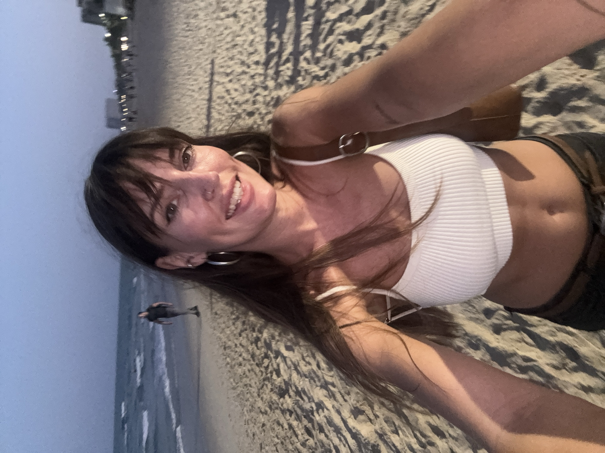 The image displays a selfie taken by a female subject on My Khe Beach in Đà Nẵng, Vietnam, during twilight or early evening. The subject occupies the central foreground, smiling directly at the camera, with long dark hair, bangs, a nose piercing, and silver hoop earrings. She is wearing a white ribbed crop top and dark-colored bottoms, with a brown bag strap visible over her right shoulder. Her right arm is extended to hold the camera. In the left mid-ground, a blurred male figure in dark attire walks away from the camera along the wet sand near the ocean, which shows small waves. The right mid-ground and background feature a sandy beach extending towards a distant, blurred cityscape characterized by illuminated buildings and indistinct human figures, indicating a developed coastline. The environment is dimly lit by the fading natural light and artificial sources from the distant city.