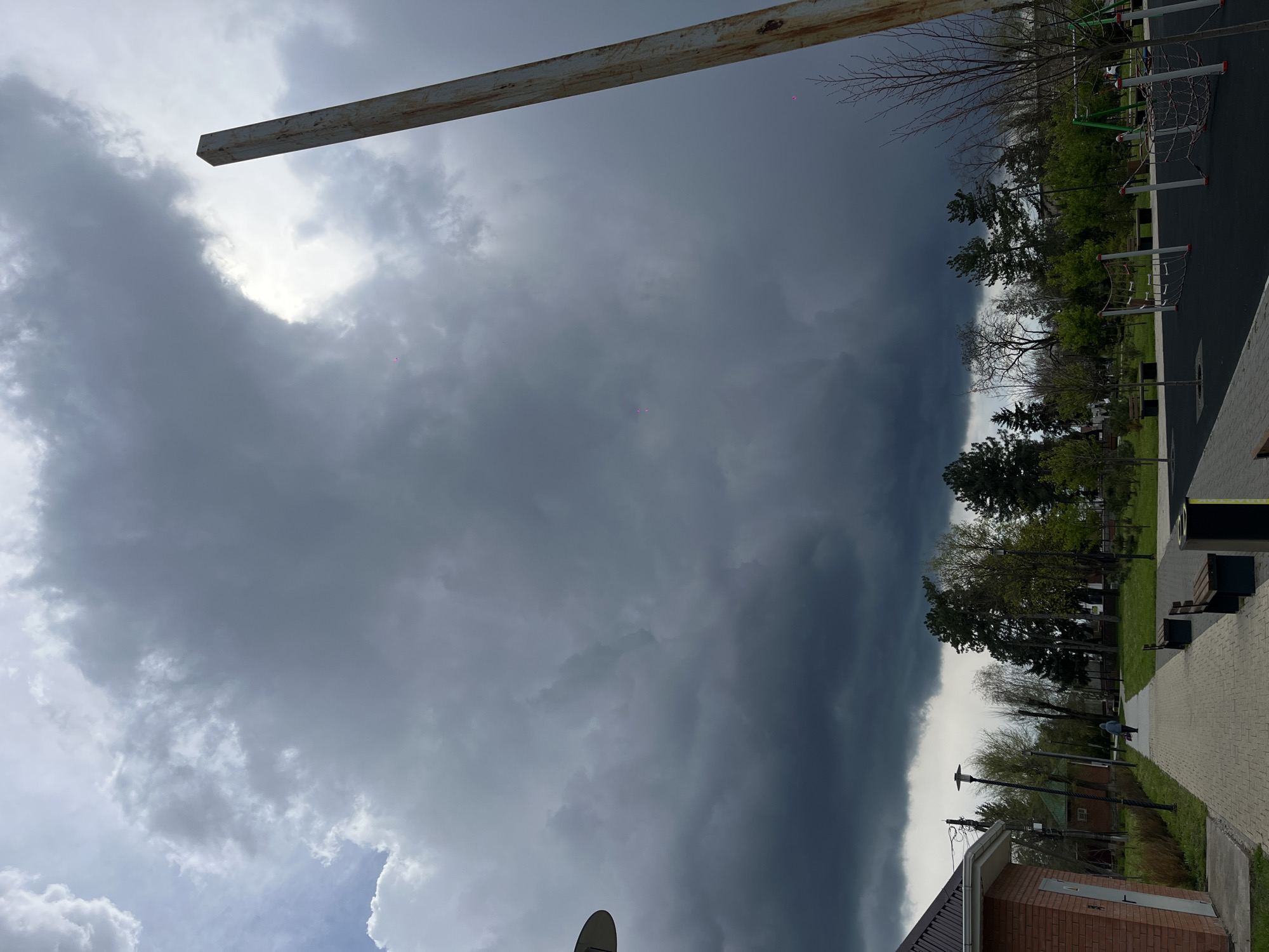 The image depicts a park in Novopokrovskaya, Russia under a stormy sky. A dark, ominous cloud formation dominates the sky, partially obscuring the sun. In the foreground, a playground area with various climbing structures and a soft, dark-colored surface is visible. Beyond the playground, trees line the park, their branches bare in some areas and showing early signs of foliage in others. A paved pathway runs along the right side of the frame, with benches and trash receptacles positioned alongside it. In the lower right corner, a portion of a brick building is visible, along with a streetlight. A metal beam is visible across the upper portion of the sky.