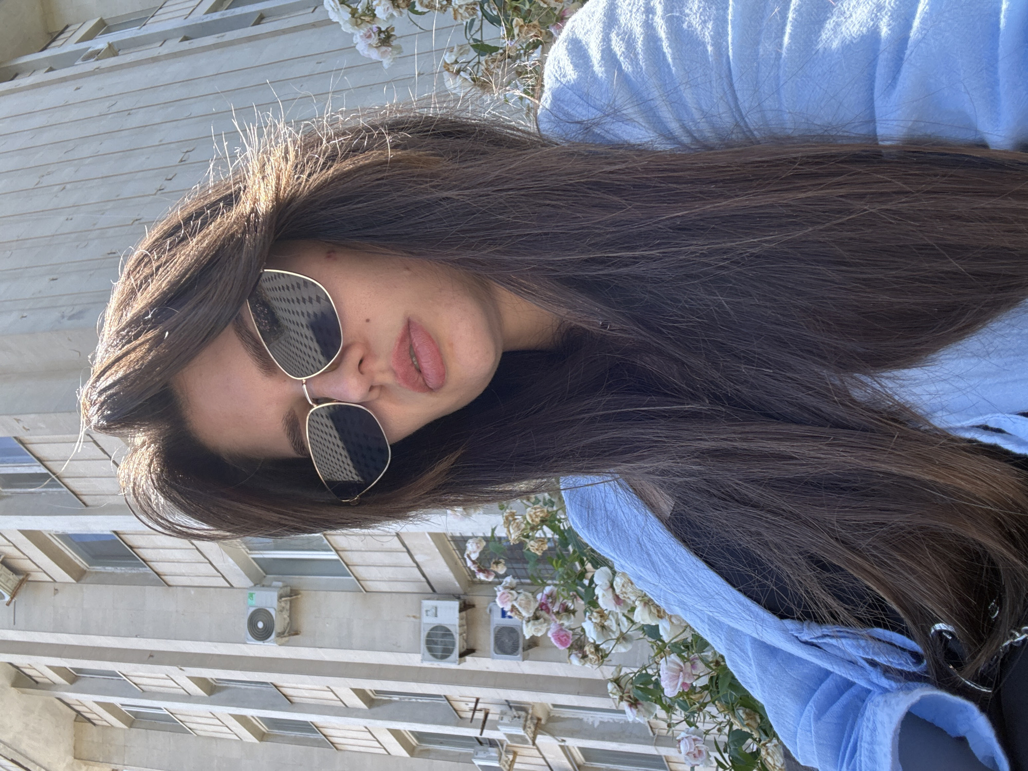 A young woman with long, dark brown hair and light skin is captured in a close-up, selfie-style photograph. She is wearing oversized, aviator-style sunglasses with gold frames. Her lips are slightly parted, revealing a hint of her teeth. She is wearing a light blue collared shirt, which is open, revealing a dark top underneath.
The background is a building with multiple windows and air conditioning units mounted on the exterior. There are also clusters of pink and white flowers with green leaves growing near the building. The lighting suggests it is daytime, with sunlight highlighting the woman's hair. The overall tone is casual and relaxed. No text is visible in the image.