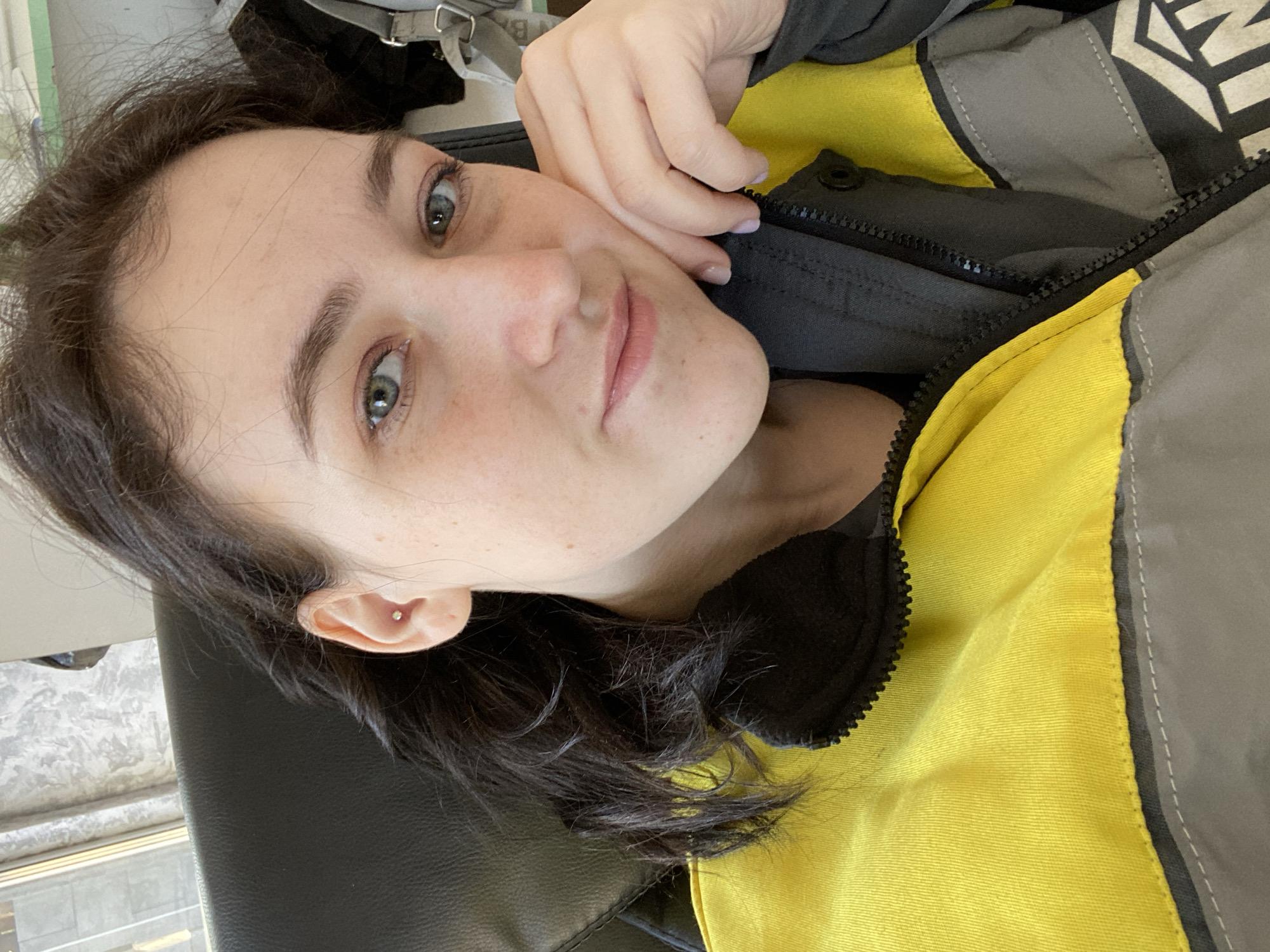 The image is a close-up, upward-angled selfie of a young woman, likely taken indoors. She has dark hair and light-colored eyes, appearing green or blue-gray, with fair skin and visible freckles across her nose and cheeks. Her expression is calm and she looks directly into the camera. A small stud piercing is visible in her right ear's tragus.
She is wearing a vibrant jacket primarily in bright yellow, with contrasting sections of gray and black. A prominent black zipper is visible down the front, and a white graphic design, possibly a partial logo or pattern featuring geometric shapes, is discernible on one of the gray sections, though it is unreadable.
Her right hand is positioned near her face, resting against her chin and the black collar of her jacket. Her fingernails are painted with a light lavender polish. She appears to be reclining or lying down on a black surface, possibly a chair or couch. In the background, blurred hints of a window or light source, along with a light-colored strap (possibly from a bag), are visible. The lighting suggests the image was taken during the daytime. The overall setting is casual, capturing a self-portrait moment. While the image itself contains no explicit location cues, the provided context indicates the location is Chaikovsky, Russia.