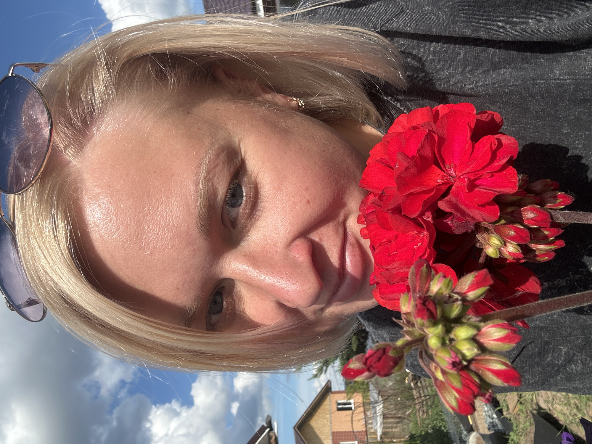 This is a close-up, upward-angle, self-portrait of a person holding a cluster of red flowers and flower buds in front of their face. The person has blonde hair and is wearing sunglasses pushed up onto their head. They are wearing a dark gray top, possibly denim. The background shows a bright blue sky with scattered white clouds, and the top of a building with a light-colored facade and a dark roof is visible on the right side. The image appears to have been taken outdoors on a sunny day. The person's expression is neutral. The focal point is the person's face and the vibrant red flowers. The composition suggests the person is taking a selfie, with the flowers framing their face. The location is identified as Chaikovsky, Russia.