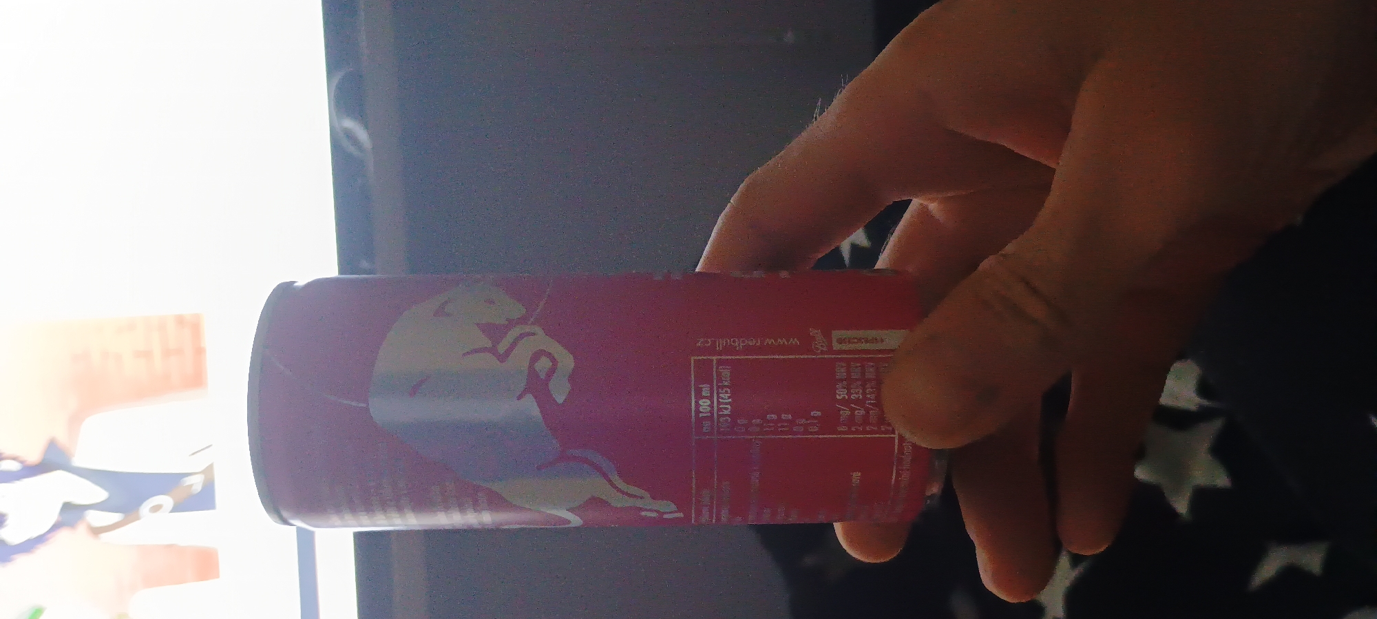 A person's hand is holding a pink Red Bull can. The can has nutritional information and the Red Bull logo printed on it. In the background, a television screen displays an anime character. The scene appears to be indoors, possibly in a living room. The image does not provide information about the specific location within Lipí, Czechia.