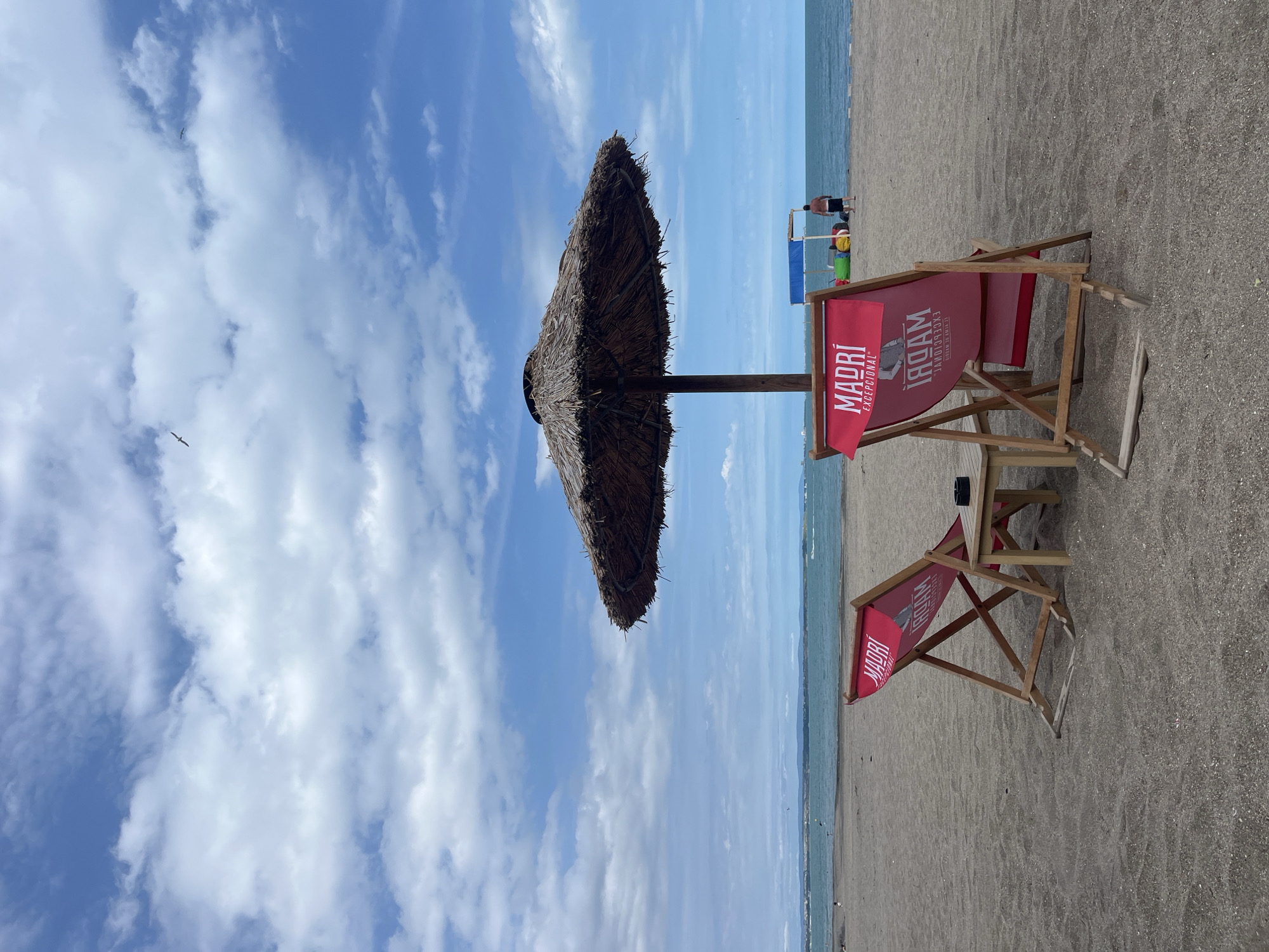 The image depicts a beach scene at the city of Burgas, Bulgaria, on a partly cloudy day. In the foreground, two empty red deck chairs with the logo "MADRÍ EXCEPCIONAL" are positioned on the sand. Between the chairs is a small wooden table. Behind the chairs, a large thatched umbrella provides shade. The sea is visible in the background, with a distant coastline. A few people are present further down the beach, near some colorful cushions and a blue canopy structure. There are also a couple of birds in flight in the sky. The overall impression is one of a quiet, relaxing day at the beach.