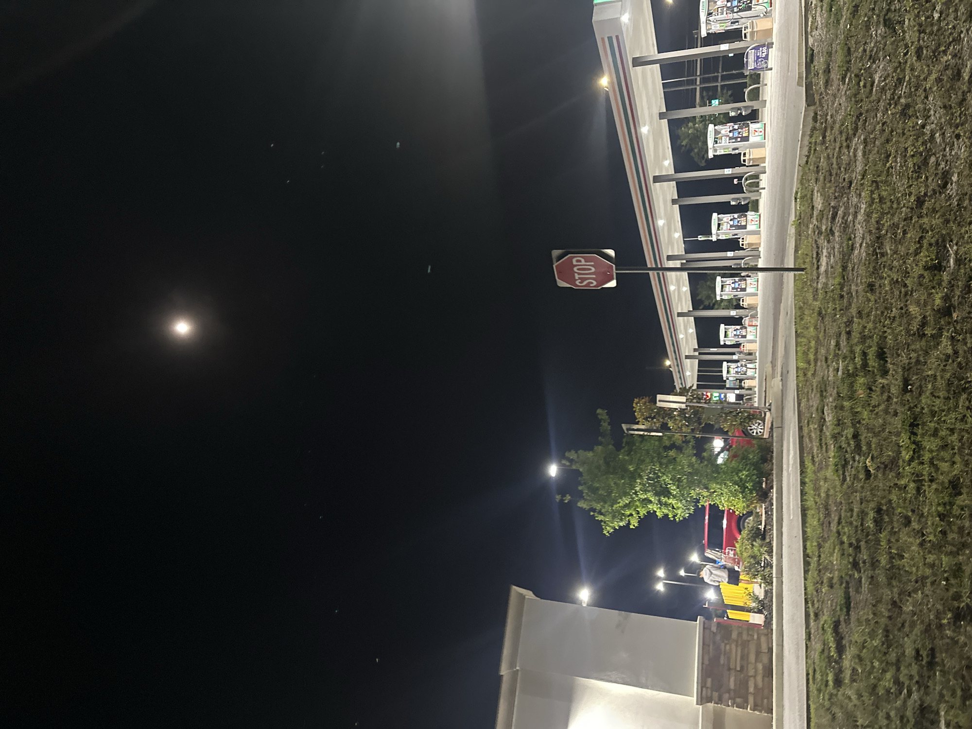 At night in Homosassa Springs, Florida, a well-lit 7-Eleven gas station is visible. Gas pumps are under a brightly lit awning. A stop sign stands near the pumps. A dark sky, with a bright moon, contrasts with the lit environment. Grass lines the right side of the scene. A partially visible building with exterior lighting and landscaping is on the left. People appear to be present, though details are indistinct. A red vehicle is parked beyond the gas station.