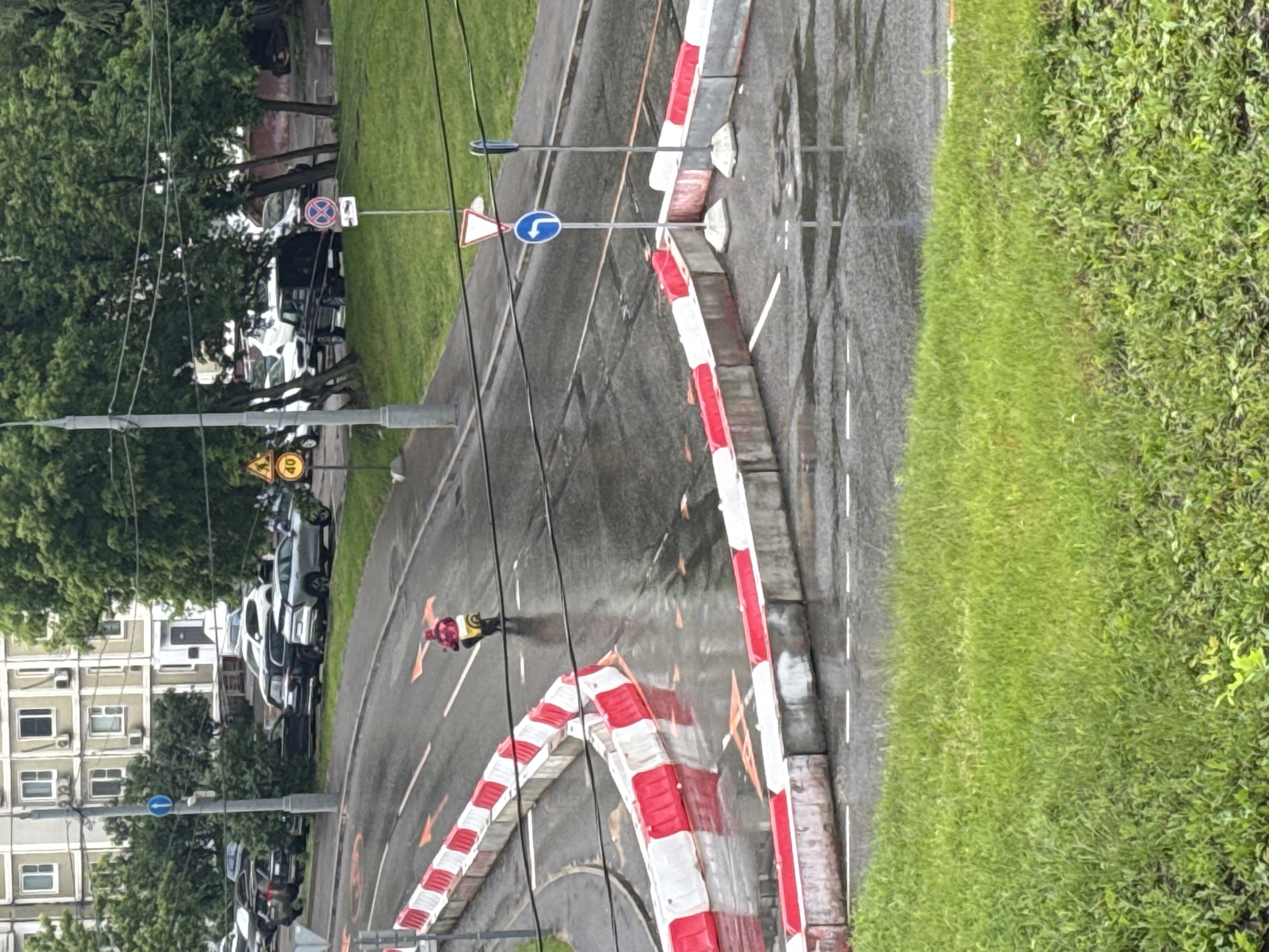 The image depicts a street scene in Moscow, Russia, after rainfall. A person on a yellow scooter, wearing a red jacket and a yellow backpack, is visible on the wet asphalt road. The road is marked with orange arrows indicating direction and is partially blocked by a series of red and white barriers. In the background, several cars are parked along the side of the road, and a multi-story building can be seen. Traffic signs, including a speed limit sign (40), a "no stopping" sign, a left turn sign, and a warning sign, are placed along the roadside. Trees and a patch of green grass are also present in the scene, contributing to the urban environment. The overall impression is one of a normal day in a city, with the recent rain adding a slickness to the road surface.