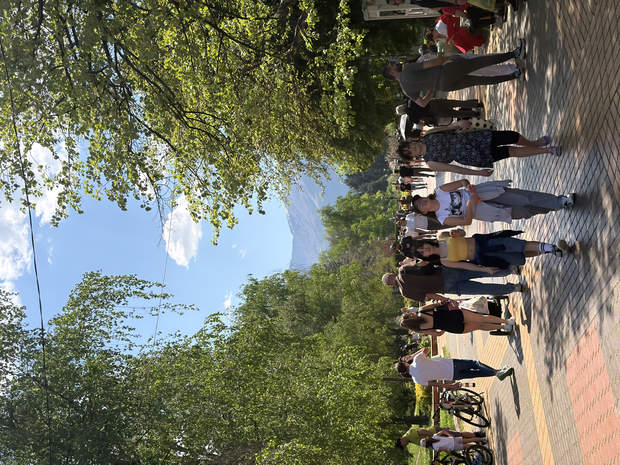 The image shows a tree-lined pedestrian walkway in Sofia, Bulgaria, crowded with people. A mountain is visible in the background. The walkway has a brick pattern, and trees line both sides, forming a canopy. The sky is blue with a few clouds. People are walking along the path, some in groups, others alone. Some people are riding bicycles, and bikes are parked alongside the walkway. Most people appear to be casually dressed.