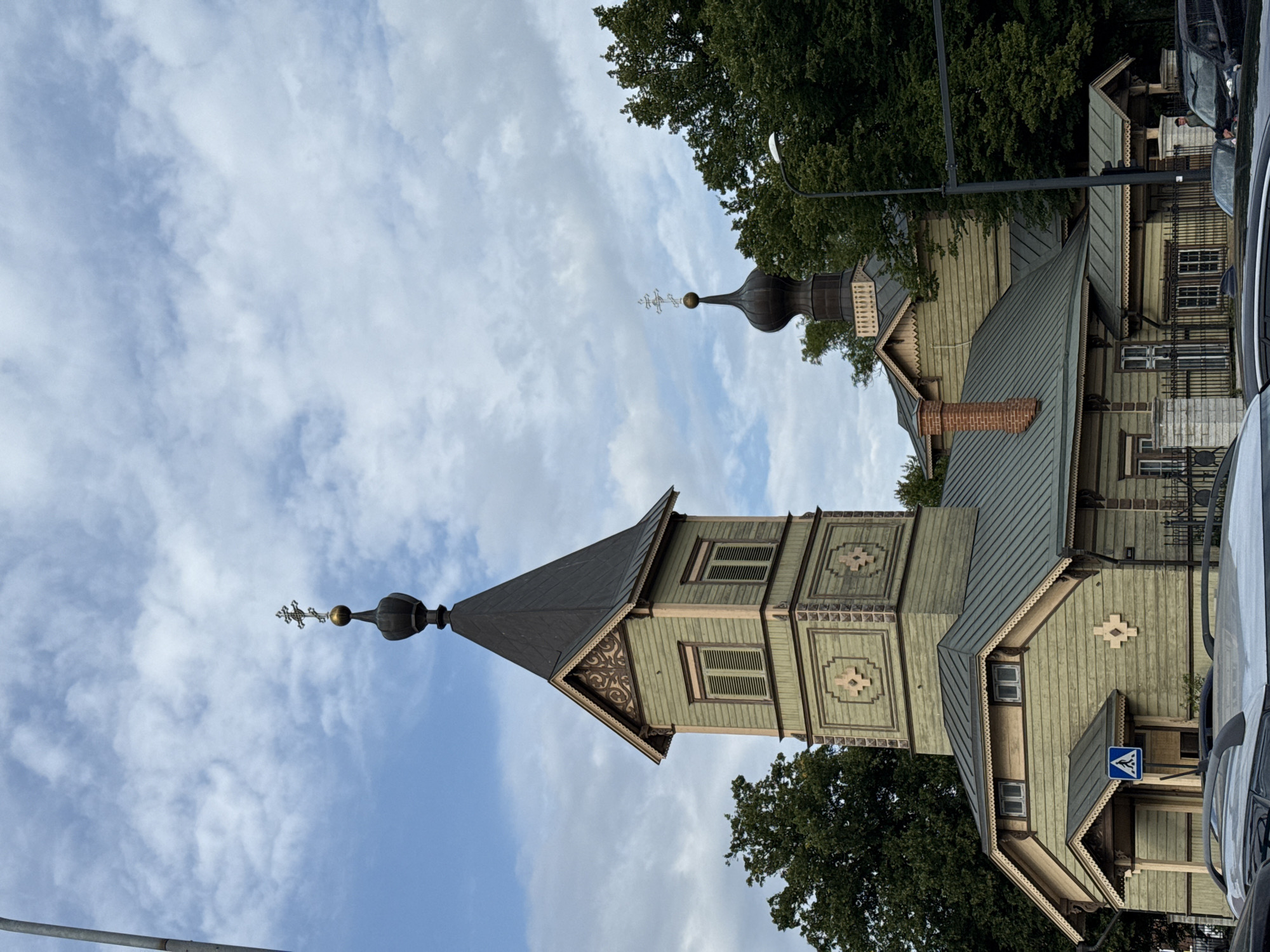 The image shows an exterior view of a light green wooden church with a dark roof. The church has a prominent bell tower with a dark spire topped with a cross, and a smaller dome with a cross behind it. The wooden facade features decorative trim and window shutters. Green trees surround the church, and a cloudy sky is visible above. In the foreground, the roof racks of several vehicles are visible, suggesting the church is located in an area accessible by car. A pedestrian crossing sign is also visible to the left of the church entrance.
