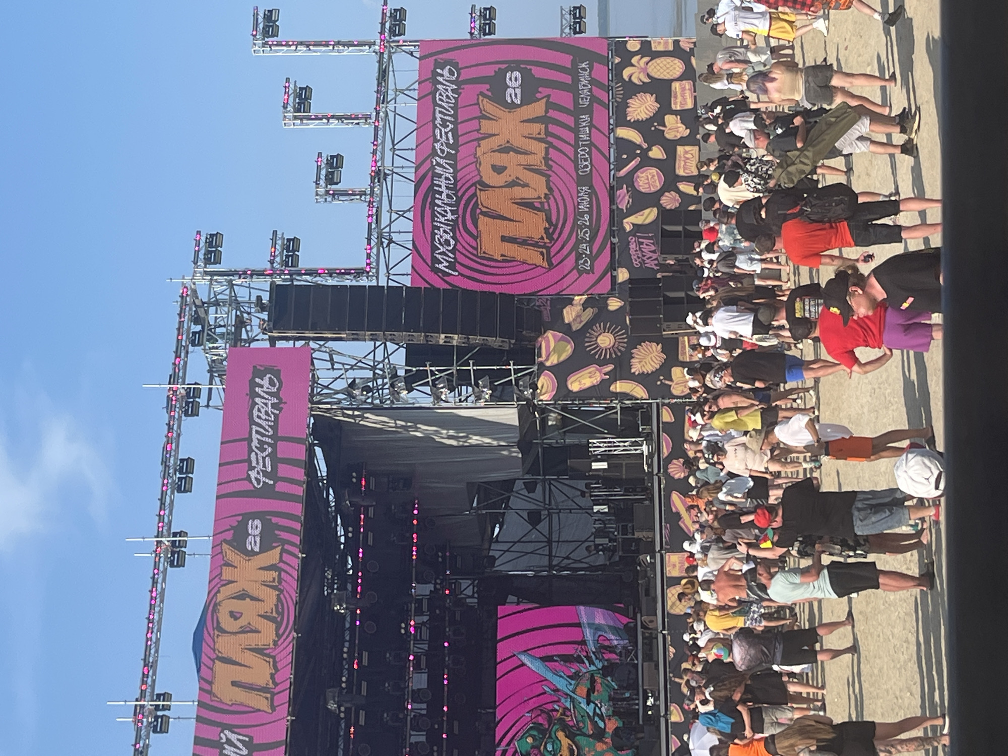 The image shows an outdoor music festival on a sandy beach, likely in Surakovo, Russia, given the Cyrillic text on the banners. A large stage with a prominent pink banner displaying "МУЗЫКАЛЬНЫЙ ФЕСТИВАЛЬ ПЛЯЖ 26" (Musical Festival Beach 26) is the central focus. The banner also features the dates "23-24-25-26 ИЮЛЯ" (July 23-26) and the location "ОЗЕРО ТИШКИ, ЧЕЛЯБИНСК" (Lake Tishki, Chelyabinsk). The stage is equipped with numerous speakers and lighting rigs.
In front of the stage, a diverse crowd of people, dressed in casual summer attire, are gathered on the beach. They are standing, talking, and some appear to be watching the stage. The ground is sandy, and the sky is clear blue. The overall atmosphere suggests a lively and festive event. The background shows a body of water, likely a lake, and the horizon. Smaller banners with fruit and ice cream motifs are also visible on the stage structure, adding to the beach theme.