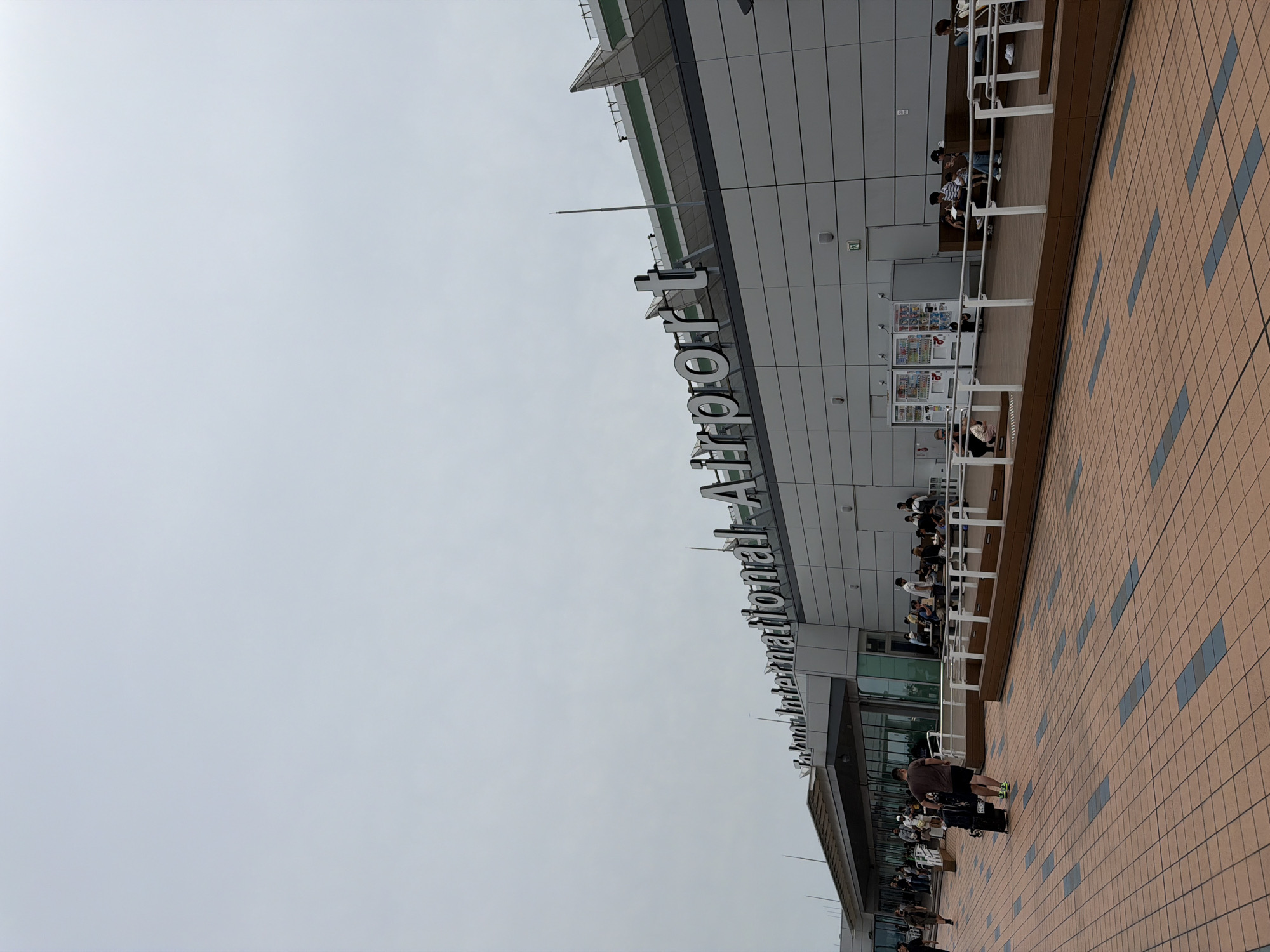 This is a wide-angle, eye-level, exterior shot of Haneda Airport in Ota, Japan, on an overcast day. The airport building is a modern, grey structure with large windows and prominent signage that reads "International Airport." The foreground consists of a reddish-brown tiled walkway with blue accents. People are visible in the midground, walking with luggage and sitting on benches outside the airport. A man is walking away from the camera, pulling a black rolling suitcase. In the distance, vending machines and more people are seen near the airport entrance. The sky is a uniform pale grey, and the overall lighting is diffused.