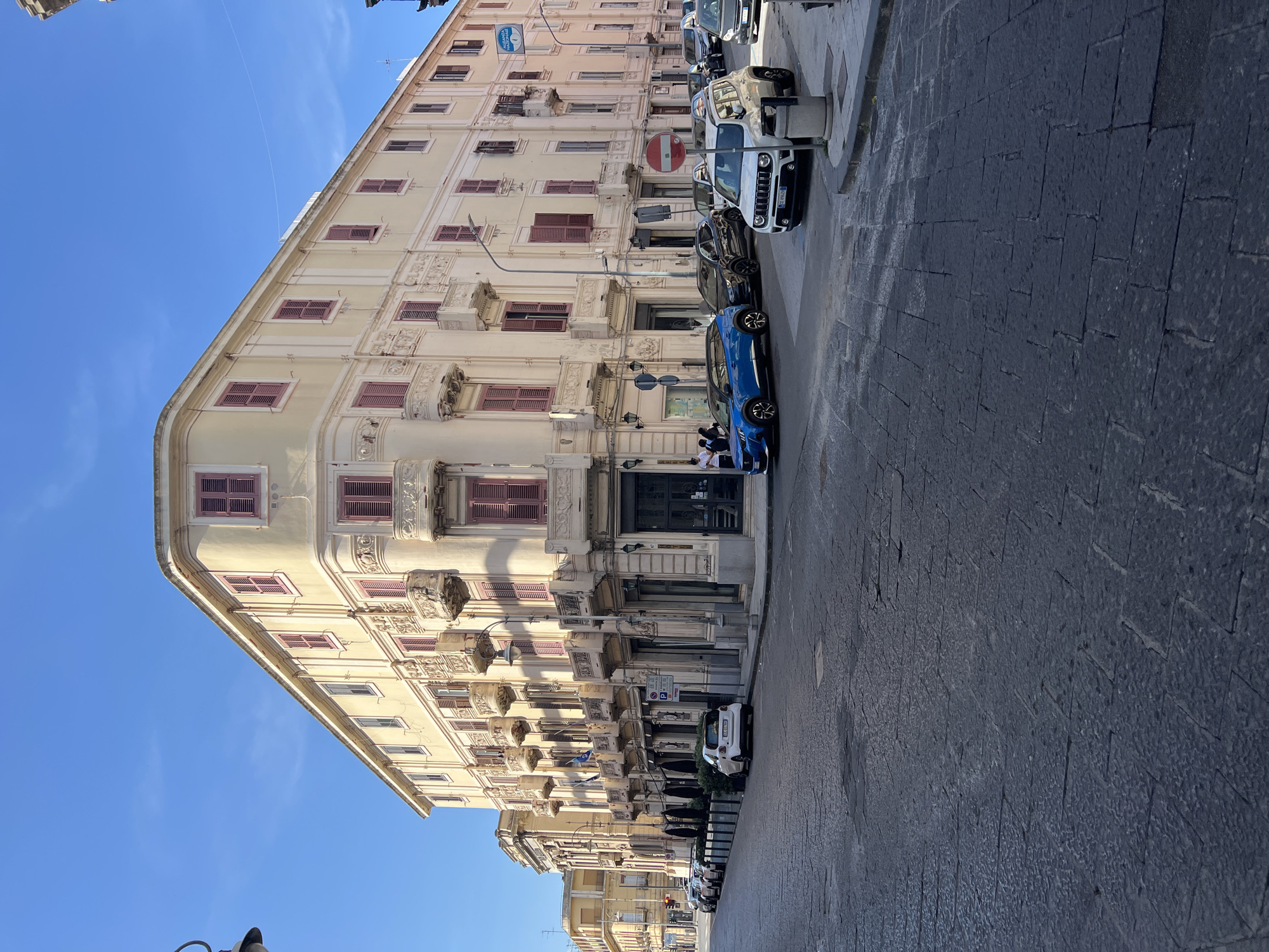 This image presents a detailed street scene in Messina, Italy, specifically focusing on the intersection involving Via Cesare Battisti 204, which is the location of the prominent corner building, the Palazzo Tremi. The central subject is a large, four-story building, painted in a light cream or pale yellow color, occupying the corner of the intersection. The building exhibits ornate architectural details, particularly on its second and third floors, which feature numerous decorative stone balconies with intricate carvings and balustrades. Each window on these floors is fitted with closed, reddish-pink wooden shutters. The ground floor of the building consists of commercial spaces with dark awnings or canopies and large windows. Above the ground floor on the right side of the building, a white circular sign with blue text "ALLIANZ NAZIONALE" is visible. The street is paved with dark grey, irregularly shaped cobblestones or flagstones, which appear worn. The scene is brightly lit by natural sunlight, casting distinct shadows and indicating clear weather under a vibrant, cloudless blue sky. Several vehicles are present in the frame: * A blue Peugeot 2008 (license plate GA 597XV) is positioned at the intersection in the foreground, facing right. * Behind the blue Peugeot, a black SUV is also at the intersection. * A white Jeep Renegade (license plate GF 696XJ) is parked at the corner curb on the right, facing right. * Behind the white Jeep, a dark-colored sedan is parked. * Further down the street on the left, a white Smart car (license plate GE 639XZ) is parked along the sidewalk, followed by several other parked cars, including a dark SUV and lighter-colored sedans. Street furniture includes: * Several black street lamp posts with multiple arms. * A blue "P" parking sign (indicating paid parking) near the white Smart car. * A prominent red circular "No Entry" traffic sign with a white horizontal bar, positioned at the corner near the white Jeep Renegade. * A grey cylindrical public waste bin is located near the "No Entry" sign. * Black bollards or stanchions line the edge of the sidewalk in front of the businesses on the left. A small group of approximately three to four individuals is visible on the sidewalk near the main entrance of the corner building, on the right side of the image, appearing to be standing or conversing. Another individual is partially obscured behind the white Smart car on the left.