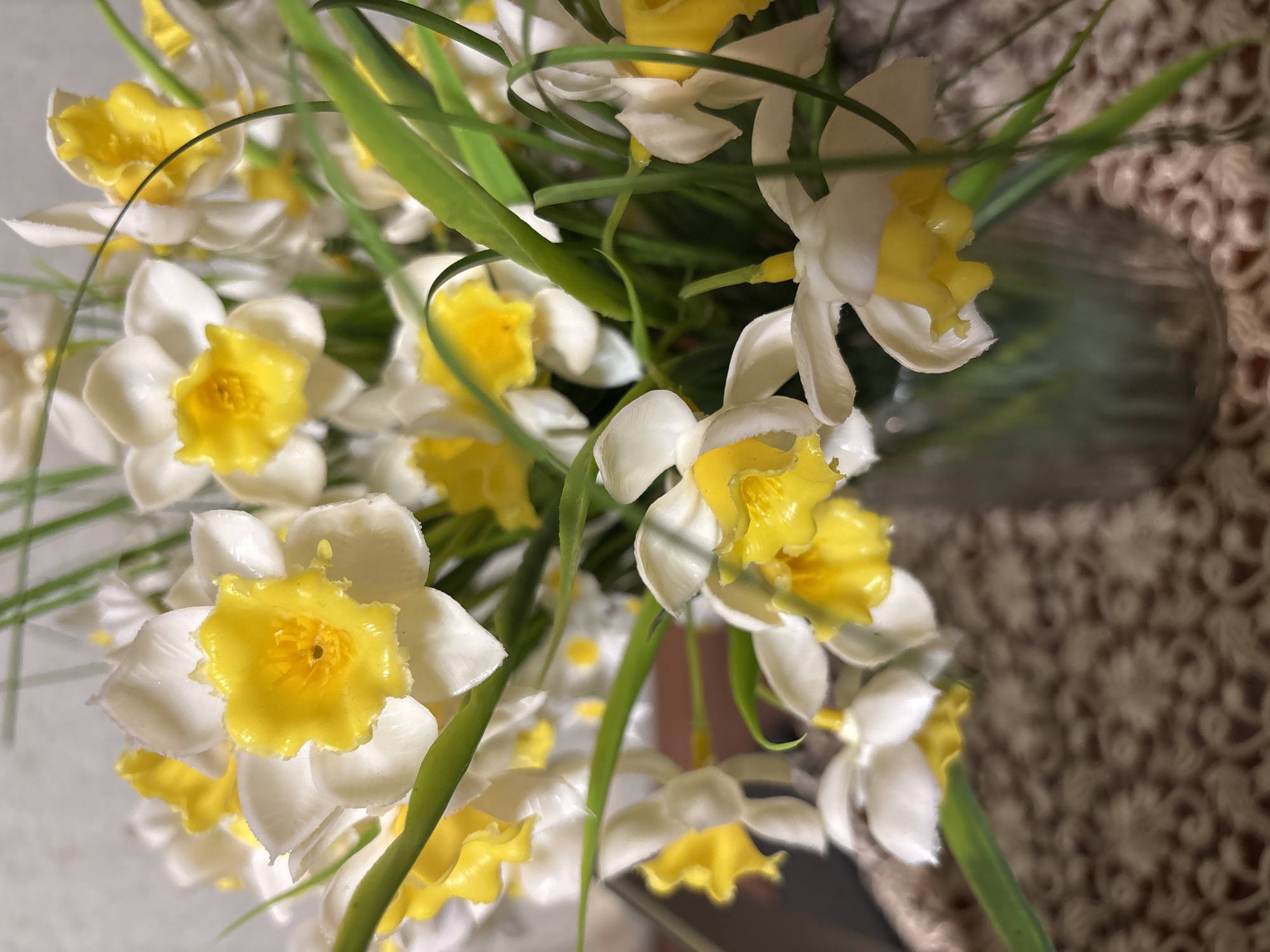 The image shows a close-up of a bouquet of white and yellow daffodils with green stems. The flowers are in focus, and the background appears to be a textured brown surface. There are no people or specific actions visible. The setting is unknown, and the information about Vagharshapat, Armenia cannot be confirmed from the image.