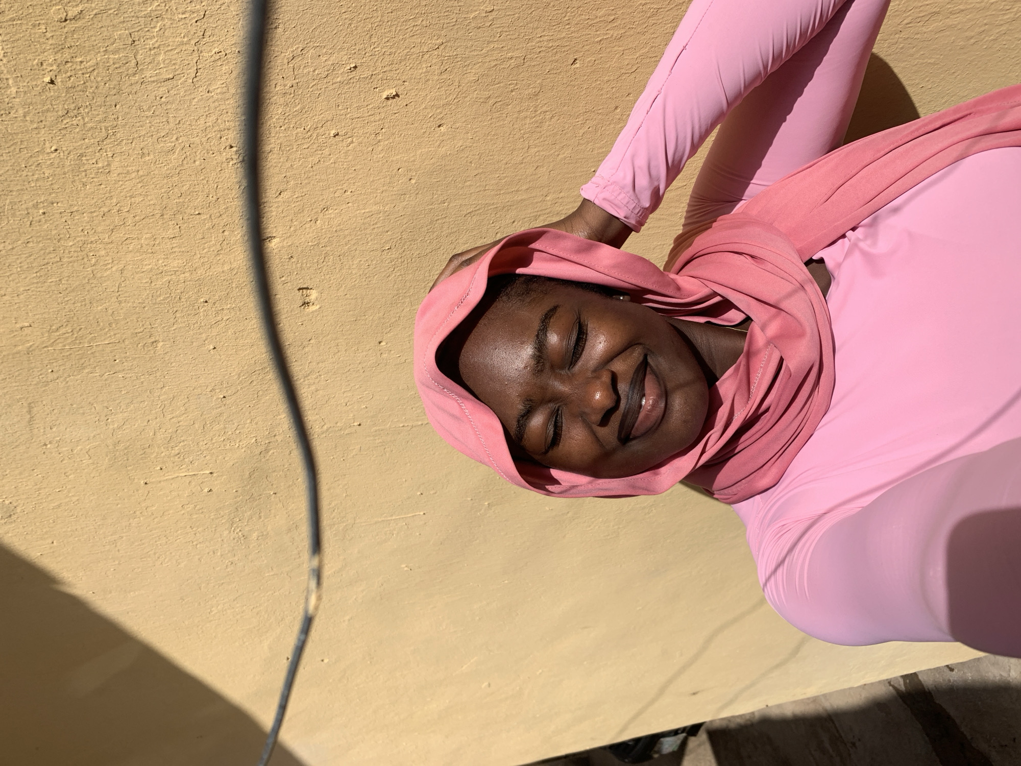 A young Black woman wearing a pink hooded garment and matching pink leggings is positioned against a yellow wall, likely outdoors in Rafin Par, Nigeria. Her eyes are closed, and she has a slight smile. A shadow from a wire or cable is cast on the wall to the left of her. The scene is brightly lit, suggesting it is daytime. There are no other observable individuals or significant objects. The overall impression is of a simple, unadorned setting.