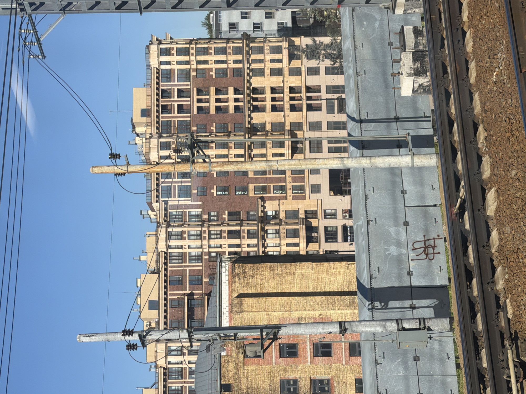 The image depicts a contemporary urban scene, likely in Moscow, Russia, viewed from a train track. Dominating the frame are several multi-story buildings with diverse architectural styles. The building in the foreground on the right is a modern, light-colored structure with many windows, partially obscured by a tall, gray utility pole. To its left, a prominent building features a façade of alternating light beige and red brick sections, with numerous windows and balconies, suggesting a residential complex. Further left, an older, taller building with a yellow brick exterior is visible, also featuring multiple windows.
In the mid-ground, a long, low, gray metal structure, possibly a noise barrier or utility enclosure, runs horizontally across the scene. It has a series of evenly spaced vertical seams and is marked with graffiti in a reddish-brown color on the right side. Several utility poles with electrical insulators and wires are interspersed throughout the image, indicating an active electrical infrastructure. The foreground shows a section of a railway track with ballast and ties, confirming the vantage point. The sky above is a clear, bright blue, suggesting fair weather. No people are visible in the image. The overall impression is of a bustling, developed urban area adjacent to a transportation corridor.