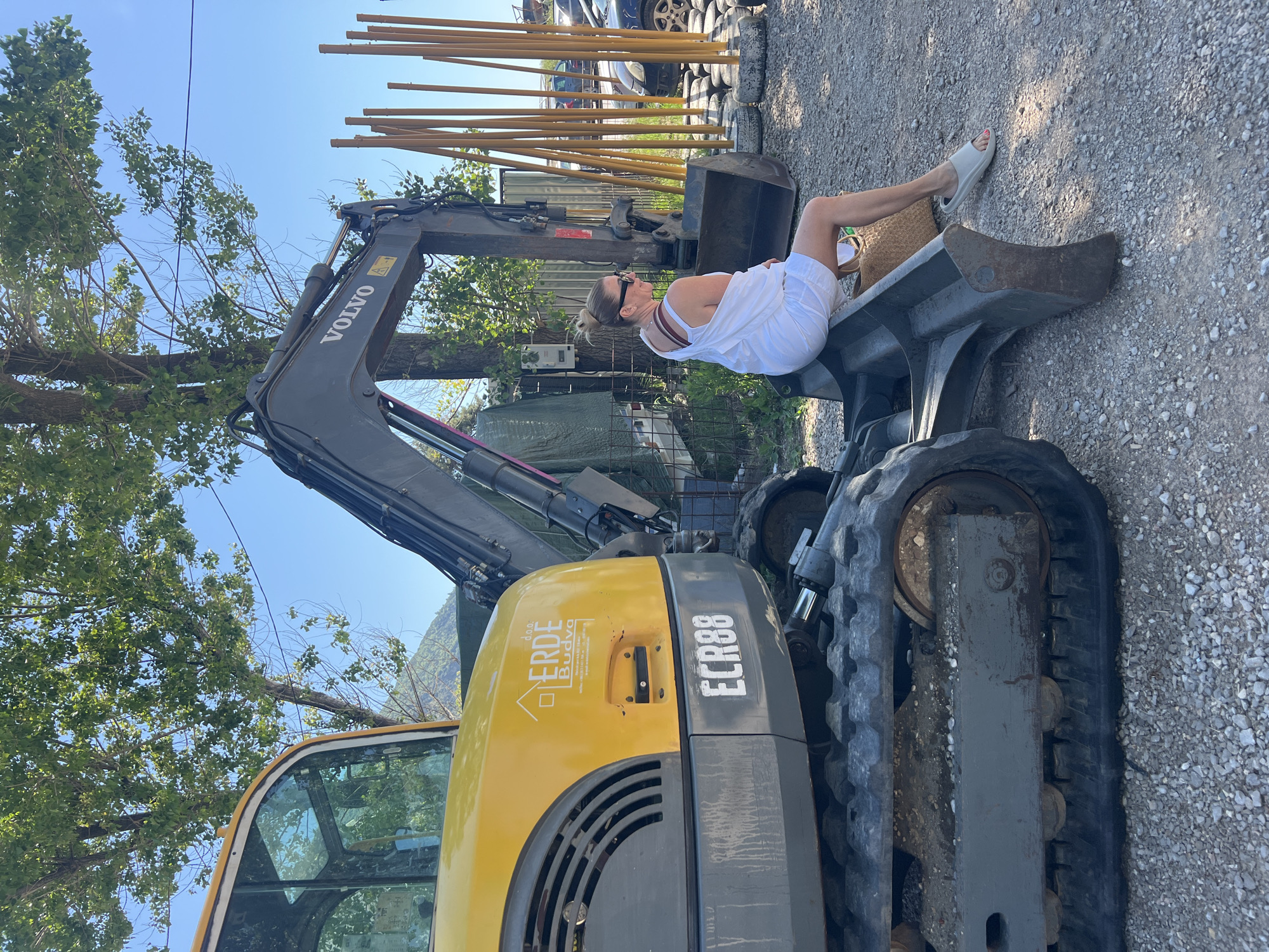 The image captures a sunny outdoor scene, likely a construction or industrial yard in Prijevor, Montenegro. A woman is casually seated on the bucket of a yellow and gray Volvo ECR88 excavator, looking towards the right side of the frame. She wears a white strappy dress, white slide sandals, and has sunglasses perched on her head with her hair tied back. A woven straw bag rests beside her on the excavator bucket.
The excavator is a prominent feature, with "VOLVO" visible on its arm and "ERDE Budva d.o.o." along with a house logo on its yellow side. The model number "ECR88" is clearly displayed on the gray rear section. The ground consists of light-colored gravel.
In the background, lush green trees are visible to the left, partially obscuring a clear blue sky. To the right, several tall yellow poles stand upright, with what appears to be a car and stacked tires beyond them, further suggesting a work or storage area. The overall impression is one of a relaxed moment amidst an active workspace under bright daylight.