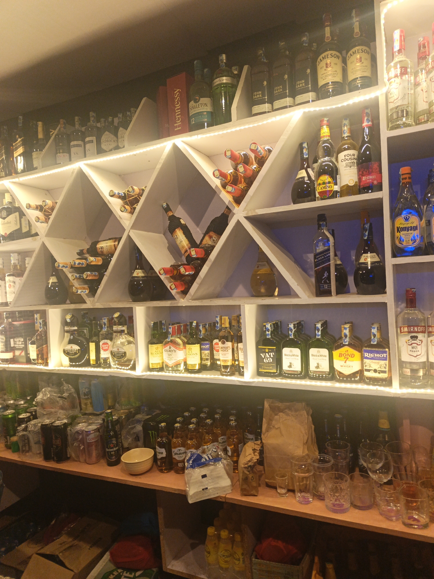 The image shows a well-stocked liquor cabinet, likely in a bar or restaurant, in Nairobi, Kenya. The shelves are filled with a variety of alcoholic beverages, including whiskey (Jameson, John Barr, Johnnie Walker Black Label), gin (Gilbey's, Hendrick's), vodka (Smirnoff), brandy (Hennessy), and various other spirits and wines. The bottles are arranged on shelves, some in triangular compartments with lights. The bottom shelf contains canned and bottled drinks, glassware, and other supplies. There are no people visible in the image.