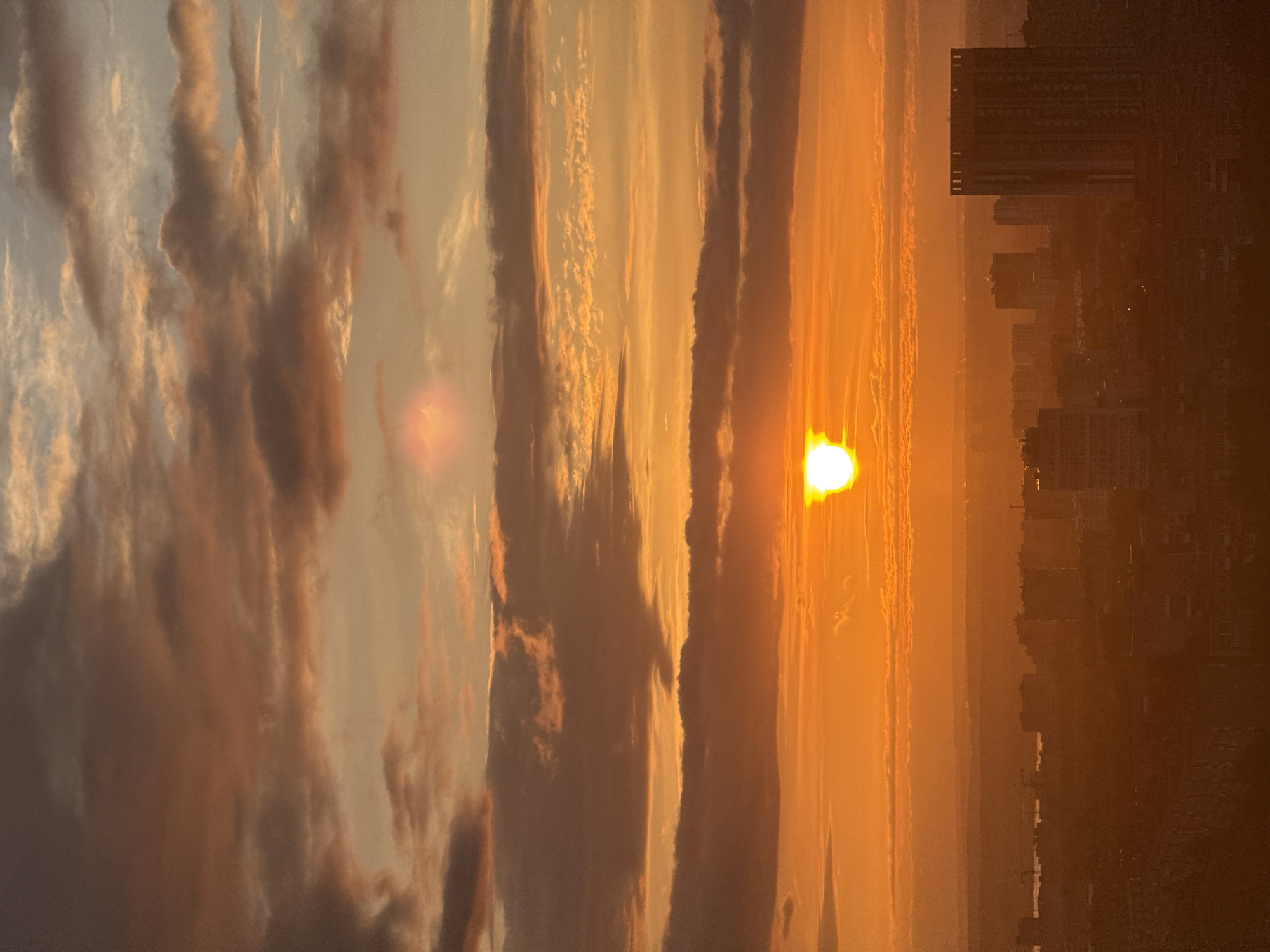 This image captures a sunset or sunrise over the city of Гамово, Russia, viewed from an elevated vantage point. The sky is largely filled with warm, intense orange and yellow hues, with the sun appearing as a bright white-yellow orb, partially obscured by clouds low on the horizon. Above the sun, several layers of clouds are visible, ranging in color from illuminated orange and gold to darker, dense grey and brown formations, exhibiting varied textures. A distinct, diffuse pink-orange light anomaly is present in the upper-middle portion of the sky. The urban landscape below is silhouetted and appears in dark brown and orange tones against the brighter sky. Numerous buildings of various heights are discernible across the city, including several prominent multi-story structures. On the far right, a tall, rectangular building with distinct features at its top stands out. In the mid-distance on the left, faint outlines of industrial structures, possibly cranes, and a distant body of water or land are visible along the horizon line. No people or specific actions are discernible within the frame.