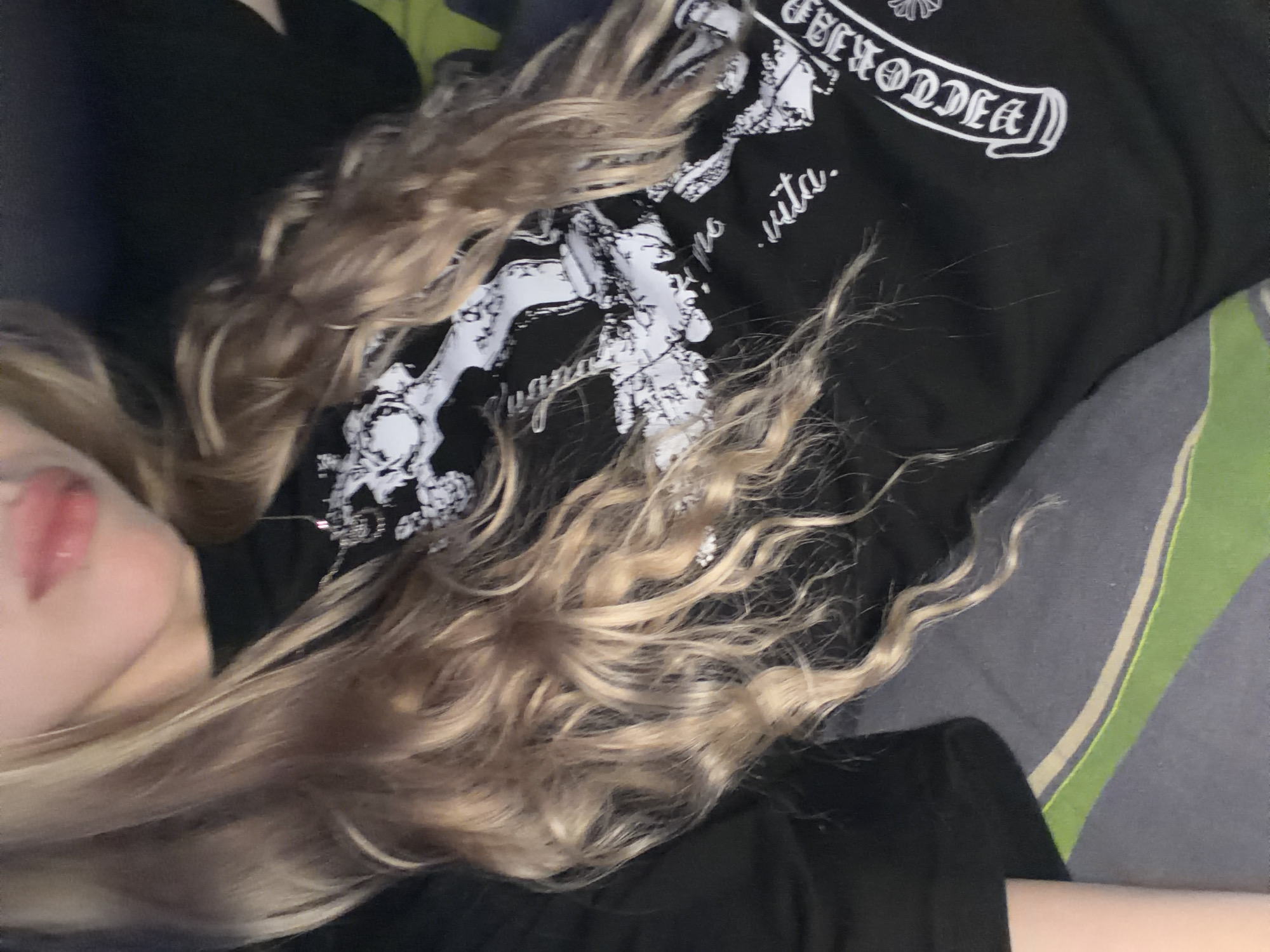 A close-up, medium shot, taken from a slightly elevated angle, shows a person lying on a bed. The person has long, wavy blonde hair that drapes over their shoulders and chest. They are wearing a black t-shirt with a white graphic print and text. The text "VICTORIA" is visible on the right side of the t-shirt in a decorative font. The person's mouth is slightly open, and their lips appear glossy. The background consists of bedding, with a dark grey duvet and a pillowcase that has horizontal stripes of grey, green, and a lighter grey. The lighting is dim, creating soft shadows. The image appears to be taken indoors in a bedroom.
