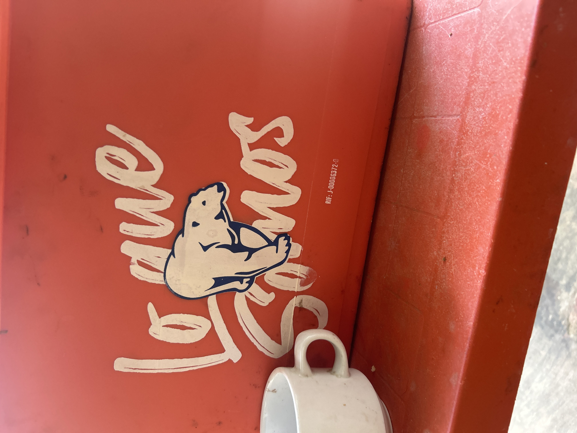 A white mug with a handle sits on a red surface, with a portion of a red cooler or container visible behind it. The cooler has white lettering that appears to say "lo que somos" and features a blue and white illustration of a polar bear. Below the illustration, there is text that reads "RIF: J-00006372-9". The red surface appears to be a table or ledge. The background is a red surface, likely part of the cooler. The scene is set outdoors, with a hint of a concrete or paved surface visible below the red ledge. There are no people present in the image. The overall setting suggests a casual, outdoor environment, possibly in Santa Ana, Venezuela, where such branding might be common on coolers or promotional items.