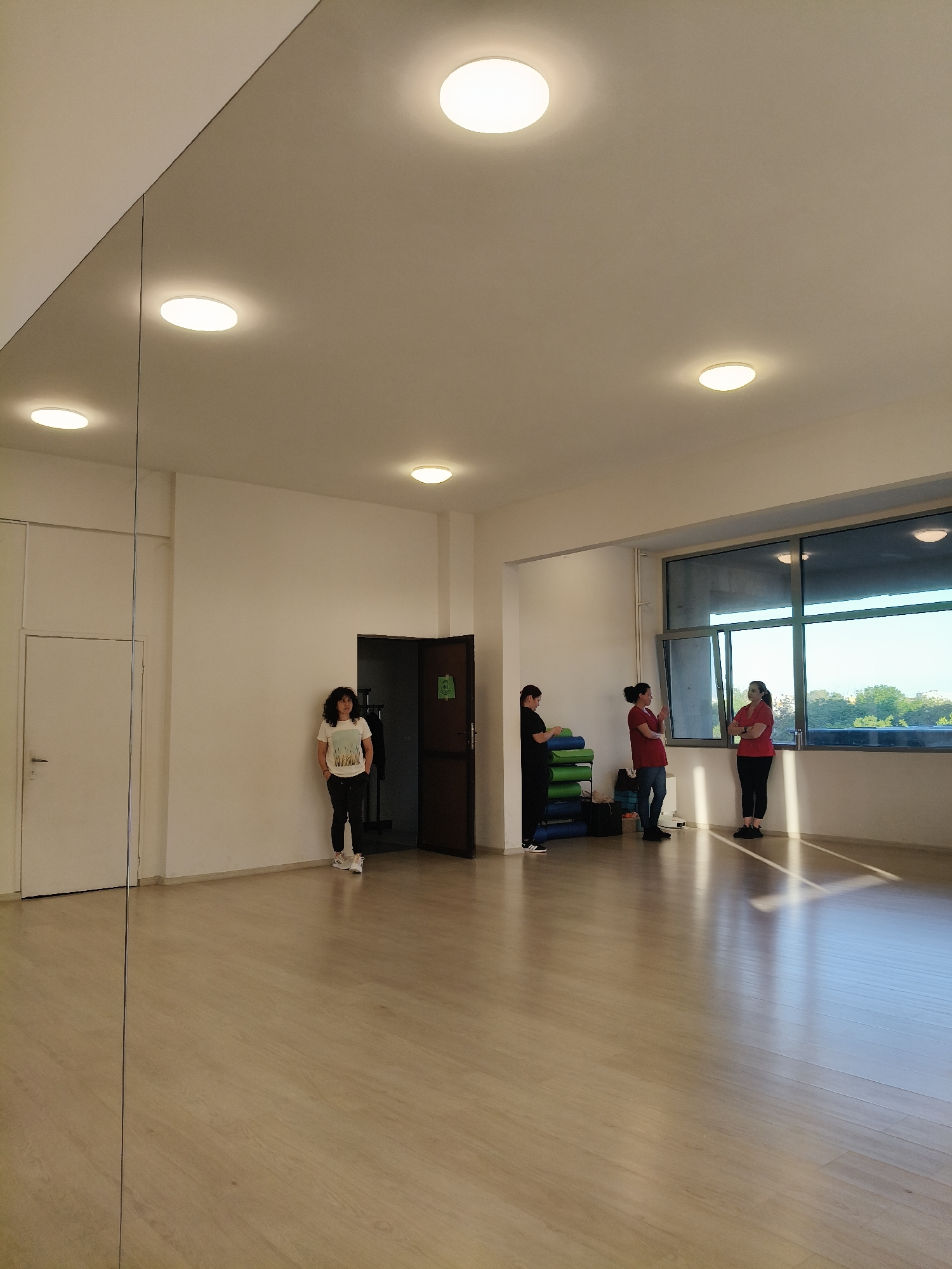 The image depicts a sparsely furnished dance studio in Plovdiv, Bulgaria, with a large mirror on one wall. The floor is a light wood-look laminate. Daylight enters through a large window. A woman stands near a closed dark brown door, with another woman and two other people near the window. Some exercise equipment is stored near the window.