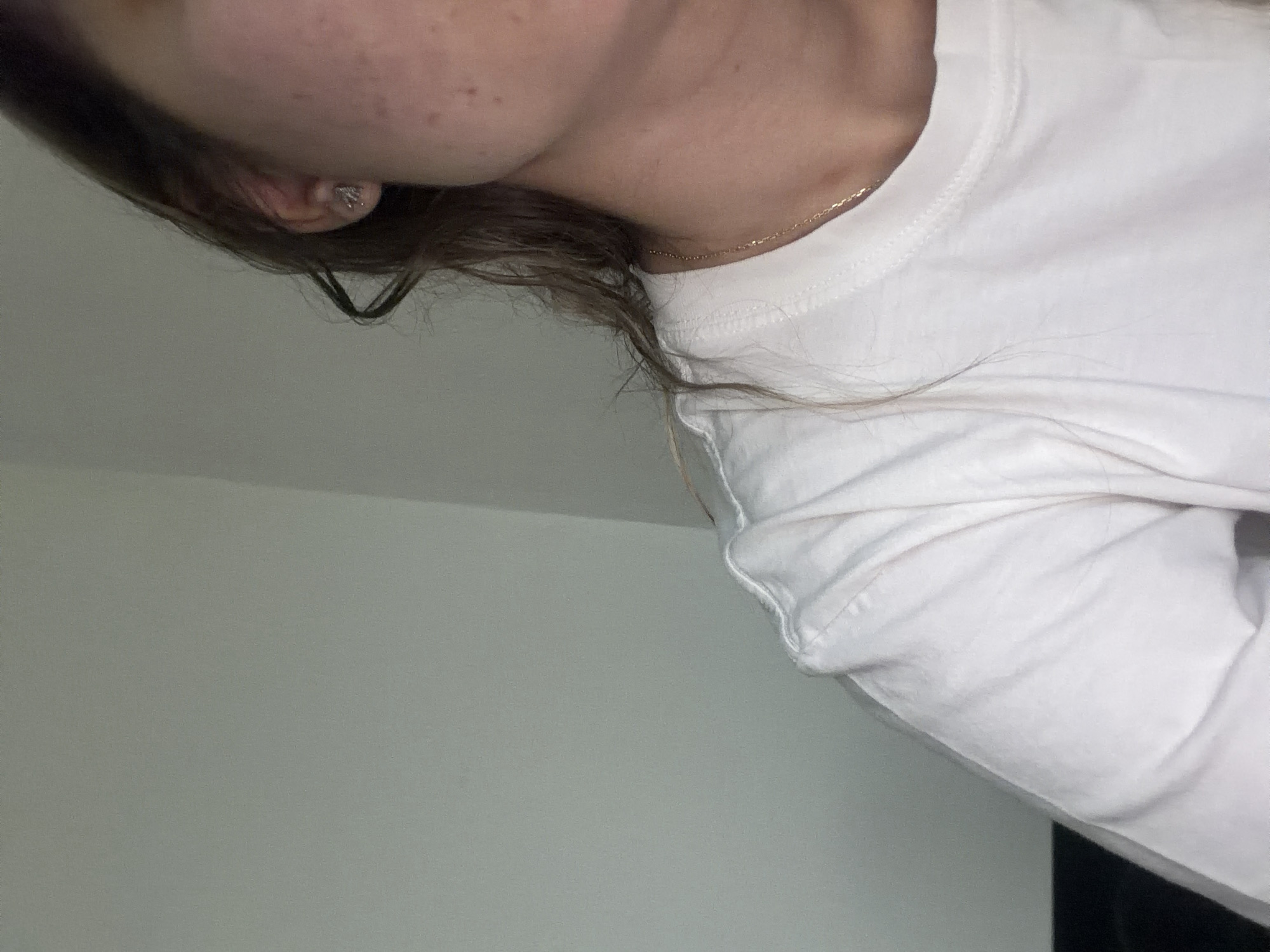 The image shows a person's head and upper body from the side, angled downwards. The person has light skin, and brown hair, and is wearing a white long-sleeved shirt and a gold necklace. A small earring is visible in the ear. The background consists of a plain wall, with a darker element in the lower right corner.
There is no evidence to determine that this image was taken in Sofia, Bulgaria.