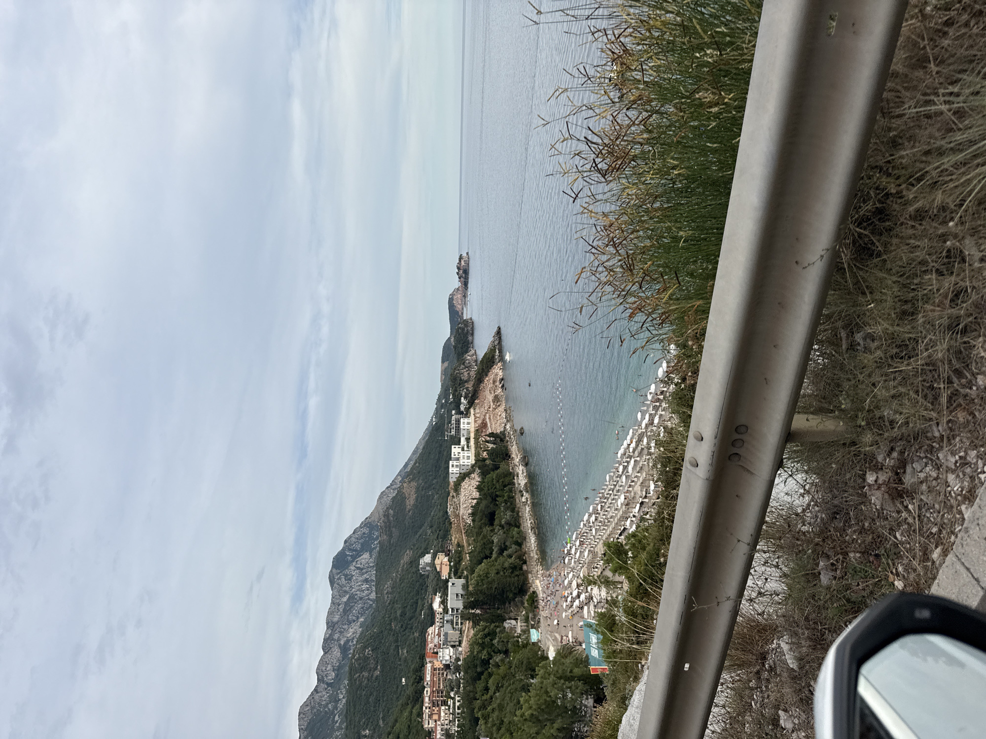 The image depicts a scenic coastal view in Rafailovići, Montenegro. The scene looks out over a calm, blue sea under an overcast sky, suggesting it might be a cloudy day or perhaps early morning or late afternoon. In the foreground, a metal guardrail runs diagonally across the frame, partially obscuring the view. To the right of the guardrail, dry, tall grasses and scrub brush are visible, indicating a natural, somewhat wild environment bordering a developed area.
The middle ground reveals a densely packed beach with rows upon rows of white beach chairs and umbrellas, indicating a popular tourist destination. People are visible on the beach, scattered amongst the chairs and walking along the shore, suggesting a leisurely daytime activity. Adjacent to the beach is a town with white buildings, nestled against the steep, green slopes of a mountain that dominates the left side of the image. The mountain is covered in dense vegetation, with some rocky outcrops visible. Further out in the sea, a small rocky island or peninsula with structures on it can be seen in the distance.
The overall impression is one of a lively beach resort town set against a dramatic natural landscape. The lighting is soft and diffused, consistent with an overcast day. No visible text is clearly decipherable in the image.
