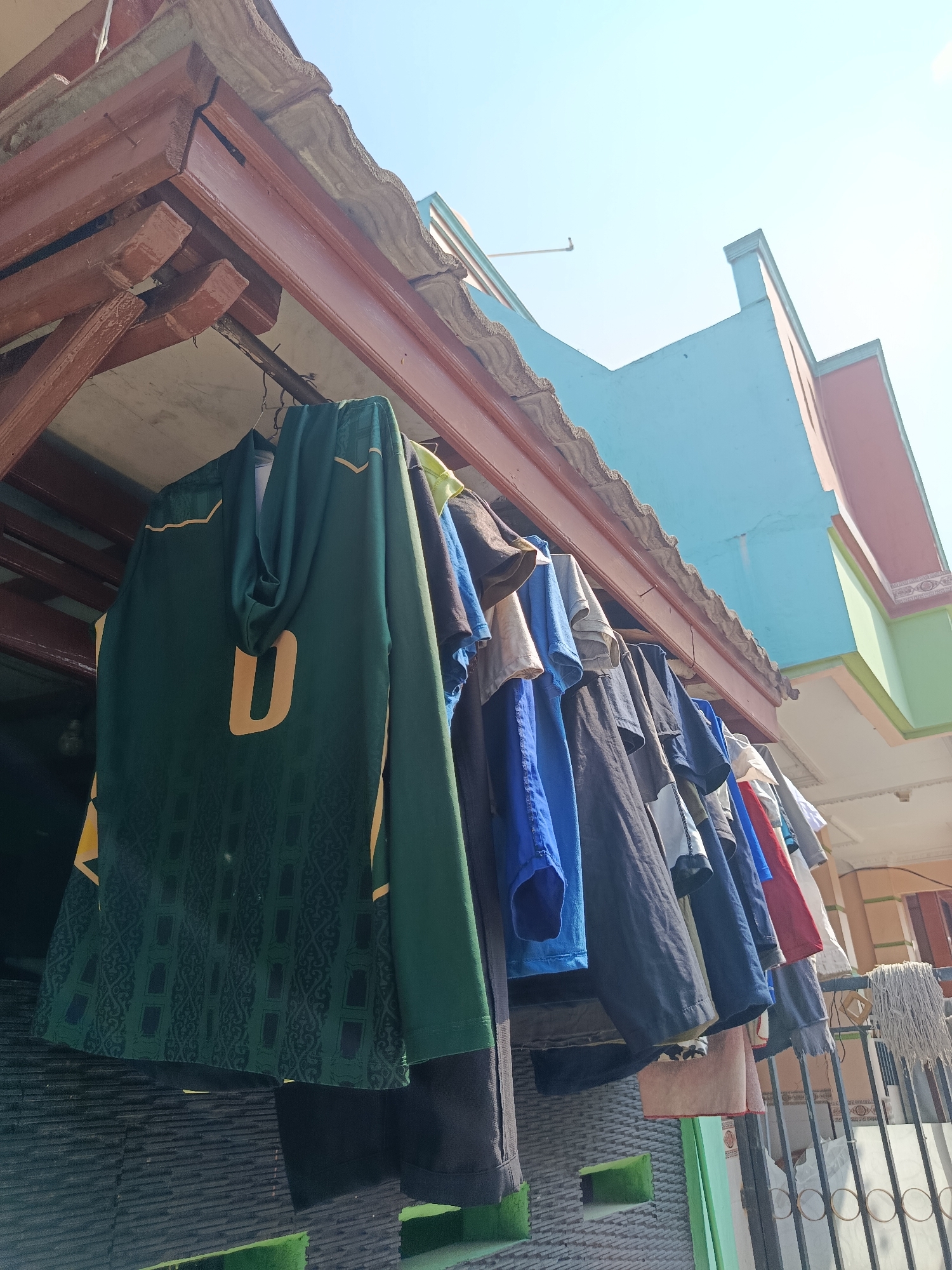 An outdoor shot captures clothes drying on a clothesline under a wooden overhang in Wanasari, Indonesia. The image focuses on a variety of shirts, including a dark green hooded shirt with a large yellow "U" on the back. Other shirts visible are blue, black, red, and white. The background shows the bright blue sky and parts of a two-story building with colorful walls in shades of blue, pink, and green. A mop and bucket are also visible to the right. The scene suggests a residential area with a focus on daily chores.