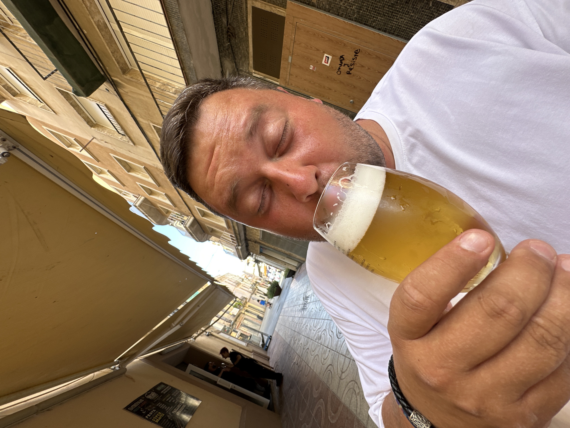 This is a close-up, low-angle selfie of a man holding a glass of beer. The man, with his eyes closed and a relaxed expression, is leaning towards the glass, seemingly about to take a sip. He is wearing a white t-shirt.
The setting appears to be an outdoor cafe or restaurant terrace in a European town. Behind the man, a narrow street is visible, lined with buildings with architectural details that suggest a Mediterranean or Southern European style. Sunlight streams down, indicating it is daytime, likely late morning or afternoon. The weather appears pleasant and sunny, with awnings providing shade over the seating area.
In the background, there are other patrons at tables and people walking down the street, suggesting a moderately busy atmosphere. One person is seen inside a doorway. A black and white menu or sign is visible on a wall near the bottom left of the image. On a wooden door in the background, some writing is visible, including "Omega Resiste". The overall mood is one of relaxation and enjoyment of a drink on a warm day.