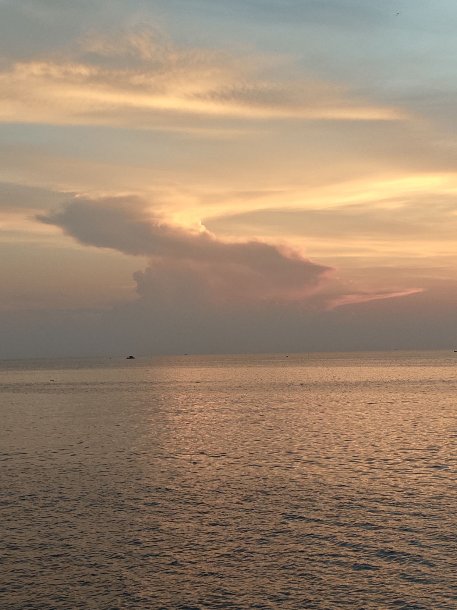 A serene seascape is captured at dusk. The sky, a gradient of light blue to peach and orange hues, is adorned with clouds. A prominent, unusually shaped cloud hovers above the water, appearing gray and pink. The sea, reflecting the colors of the sky, shimmers with golden light. The water ripples gently, suggesting a calm environment. In the distance, several small boats or ships are visible, indicating maritime activity. The location is suggested to be Lhokseumawe, Indonesia. No people are visible in the image.