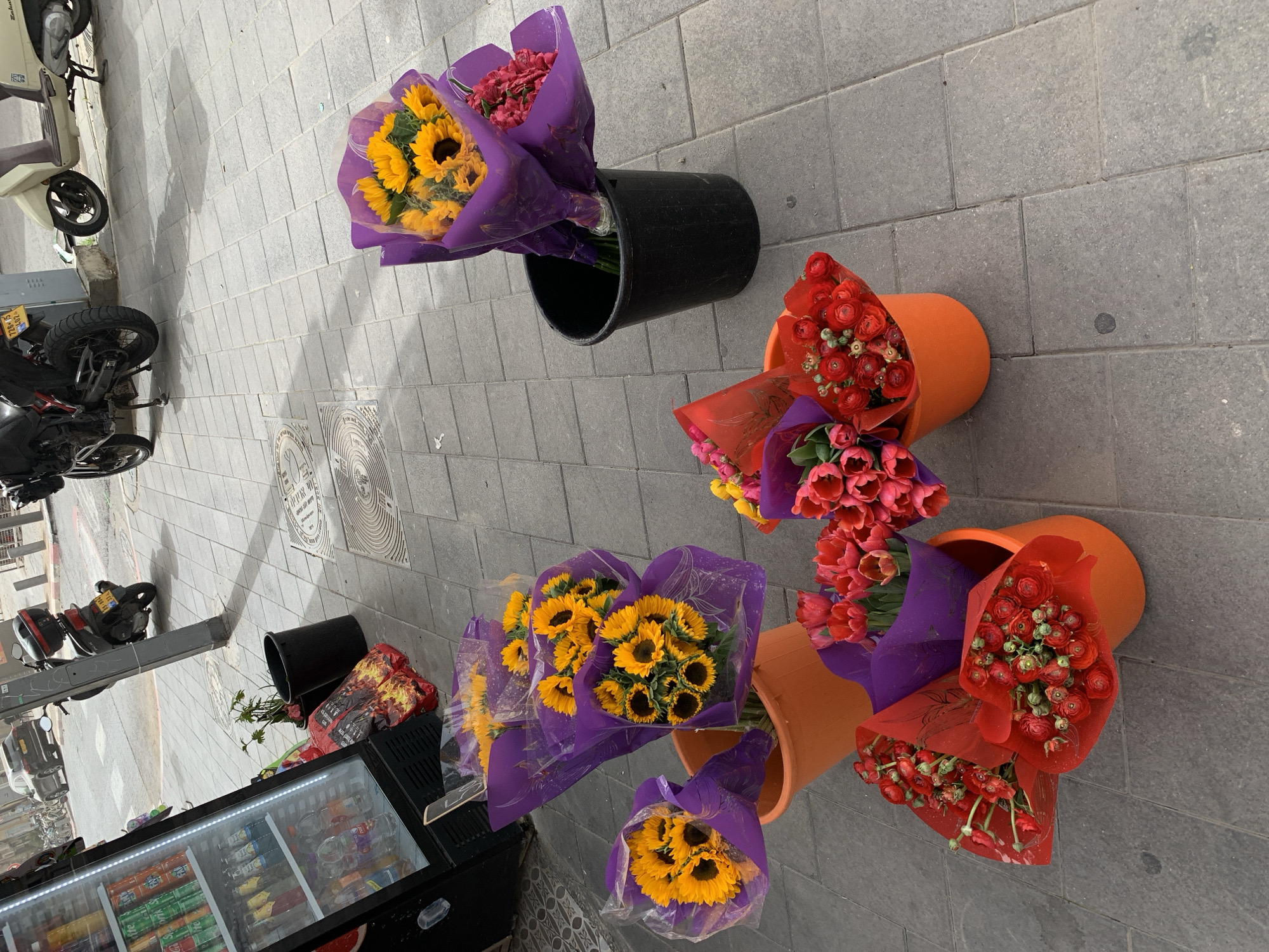 The image captures a Tel-Aviv street scene, likely a temporary flower stand on the sidewalk. Several bouquets of flowers, including sunflowers and red flowers (possibly ranunculus or tulips), are arranged in buckets. Some buckets are black plastic, while others are terracotta-colored. The flowers are wrapped in clear plastic and purple or red paper. A beverage cooler filled with drinks stands nearby, suggesting a small store or kiosk. A parked motorcycle and scooter are visible in the background, along with the sidewalk and street. The scene is shot from an elevated angle, giving a view directly down onto the flower display.