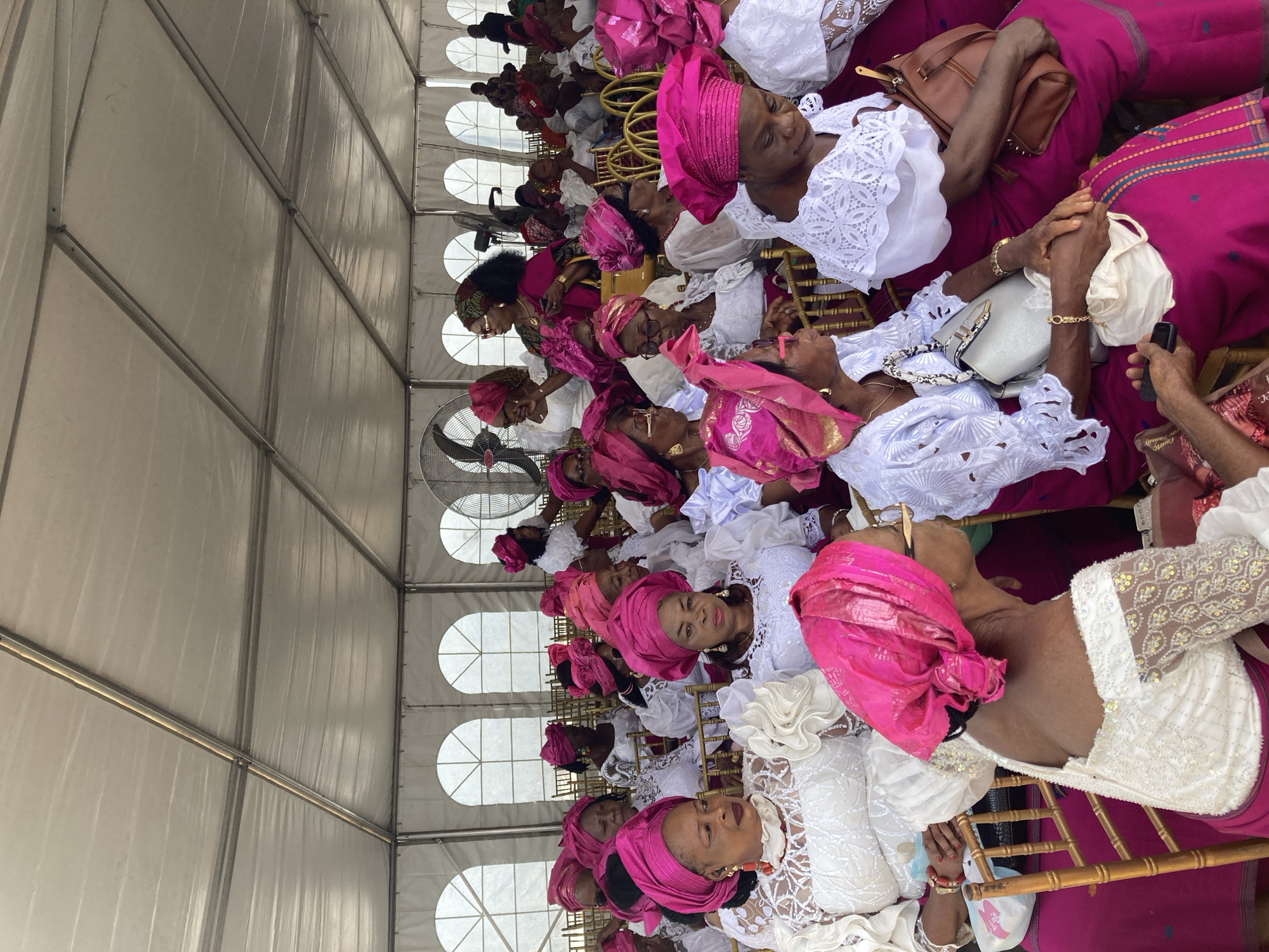 A horizontal, eye-level shot depicts a group of Black women seated in rows inside a white tent. The women are predominantly wearing white lace tops and pink wrappers and head wraps. Many are wearing sunglasses and gold jewelry. They are seated on gold chairs. The tent has a white fabric ceiling and round windows. A fan is visible at the top of the frame. The location is determined to be Nnenu, Nigeria.