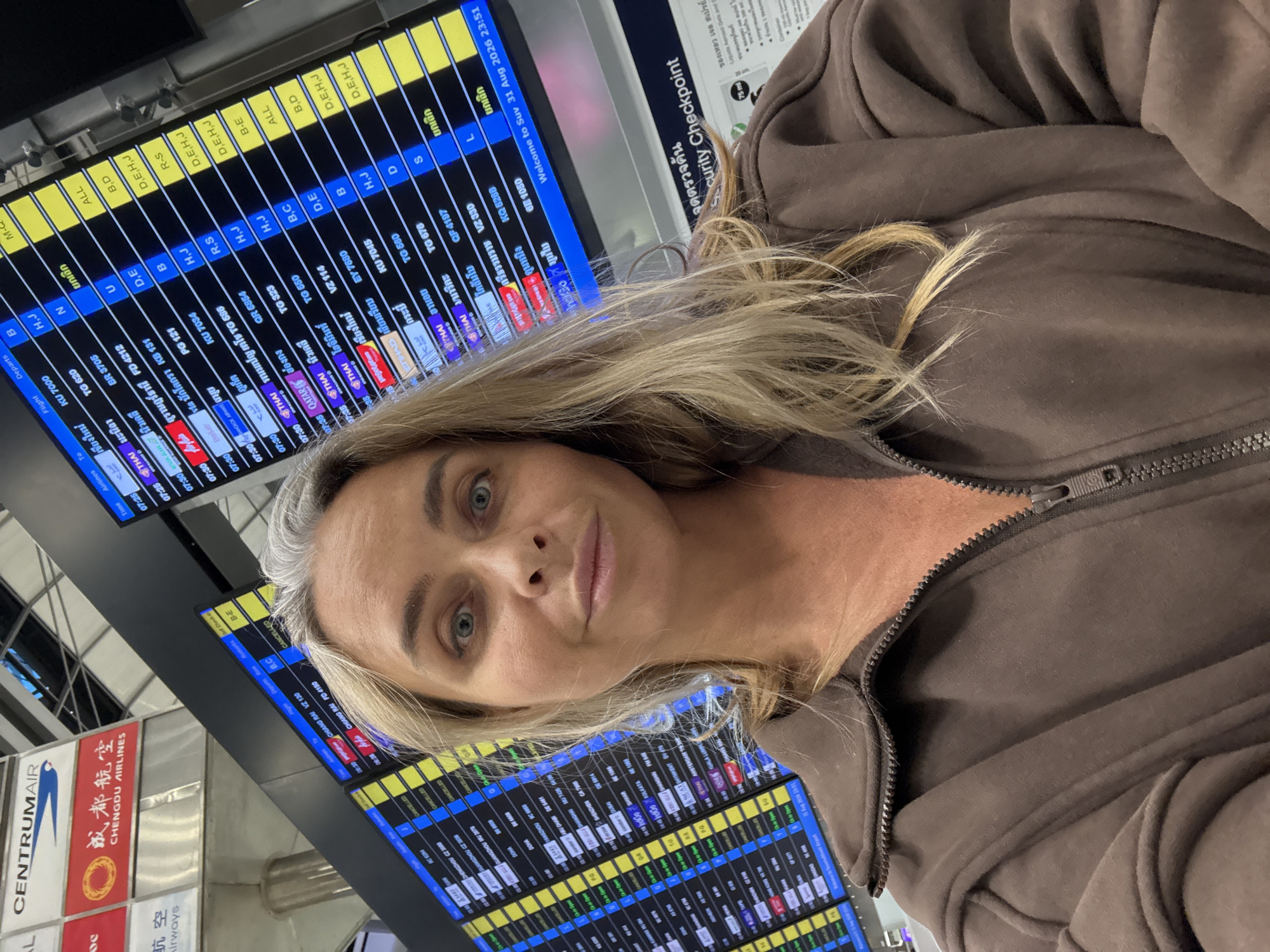 A woman with blonde hair, wearing a brown zip-up hoodie, takes a selfie in an airport. She is positioned in the foreground, with her face slightly angled towards the camera. Behind her, a large digital departure board displays flight information, including flight numbers, destinations, and times. The departure board is illuminated and appears to be operational. The background also shows airport signage and other infrastructure, suggesting a busy travel environment. The woman's expression is neutral.