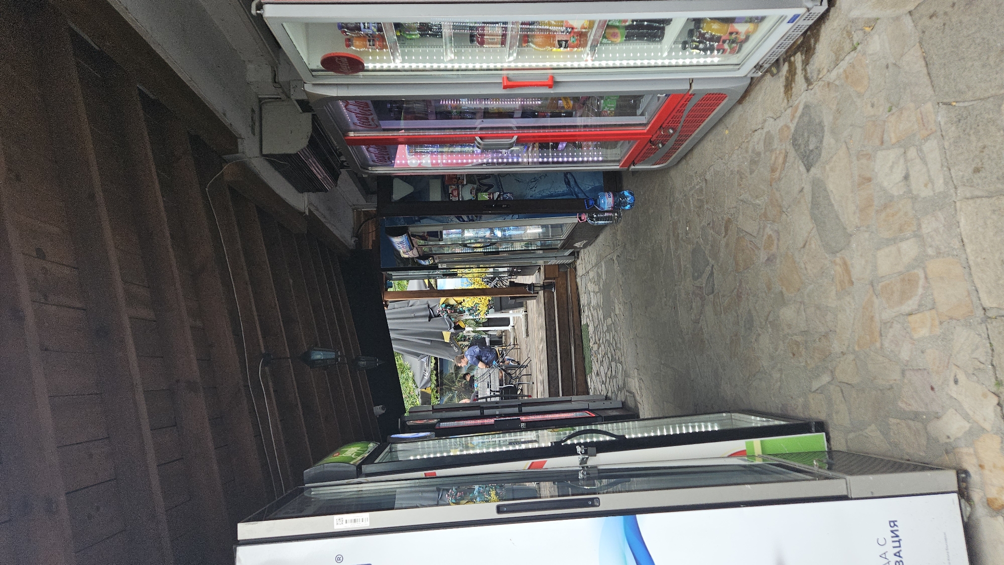 The image shows a row of refrigerated vending machines lining a stone-paved walkway in Plovdiv, Bulgaria. A wooden structure or building is on the left side of the walkway. The vending machines contain various drinks. In the distance, one or two people can be seen sitting at tables under umbrellas.