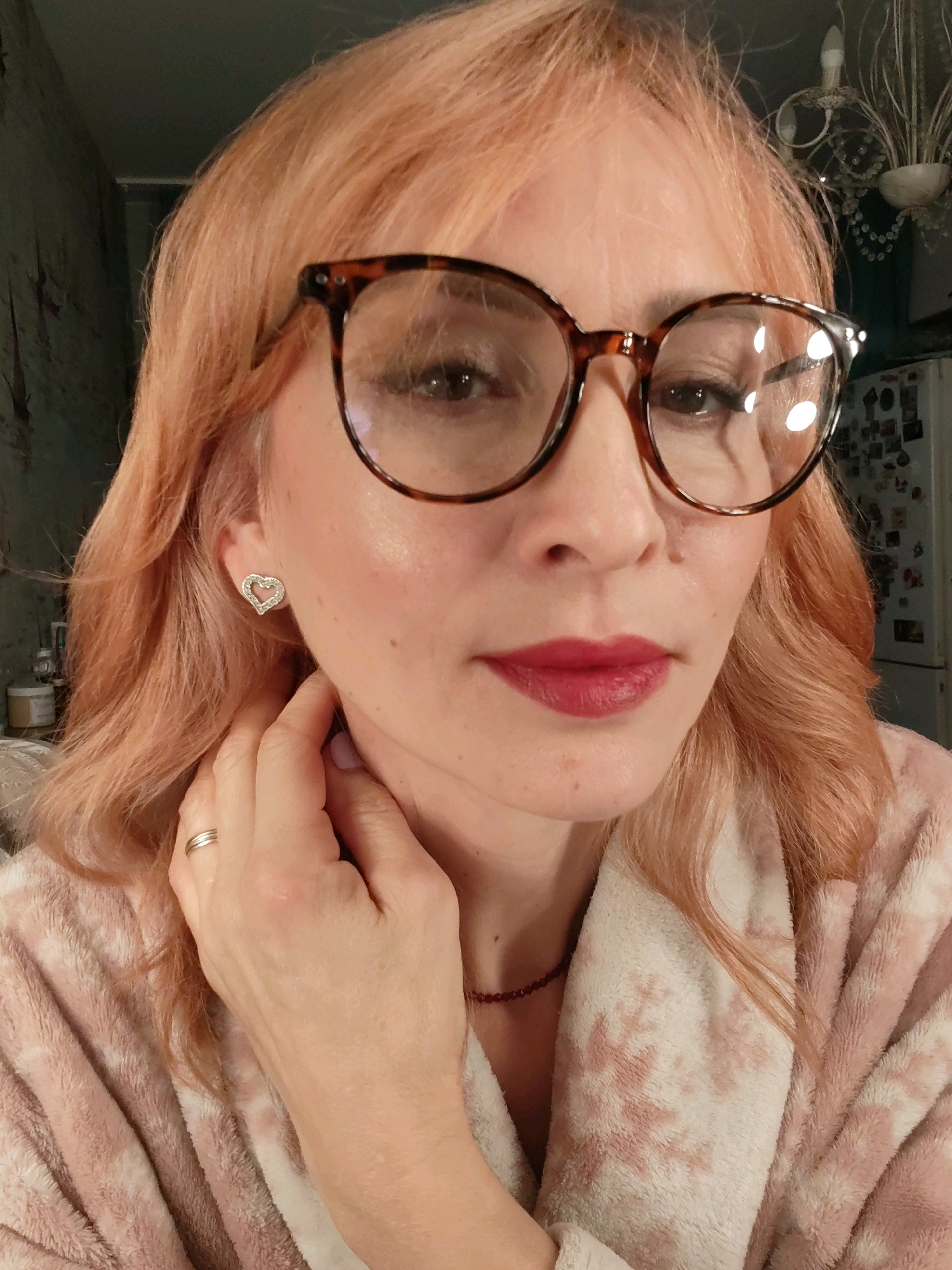 A close-up, selfie-style shot captures a woman with peachy-pink hair and large, tortoiseshell-framed glasses. She is wearing a soft, pink bathrobe with a white tie-dye pattern. Her left hand is gently touching her neck, adorned with a thin gold wedding band on her ring finger. A delicate, heart-shaped stud earring with small clear stones is visible on her left ear. Her lips are painted a deep berry red. The background appears to be an indoor setting, possibly a kitchen, with a hint of a refrigerator to the right and a somewhat rough, dark wall on the left. There is also a glimpse of a decorative chandelier hanging in the upper right corner. The image does not provide any specific information to suggest a location in Chaikovsky, Russia.