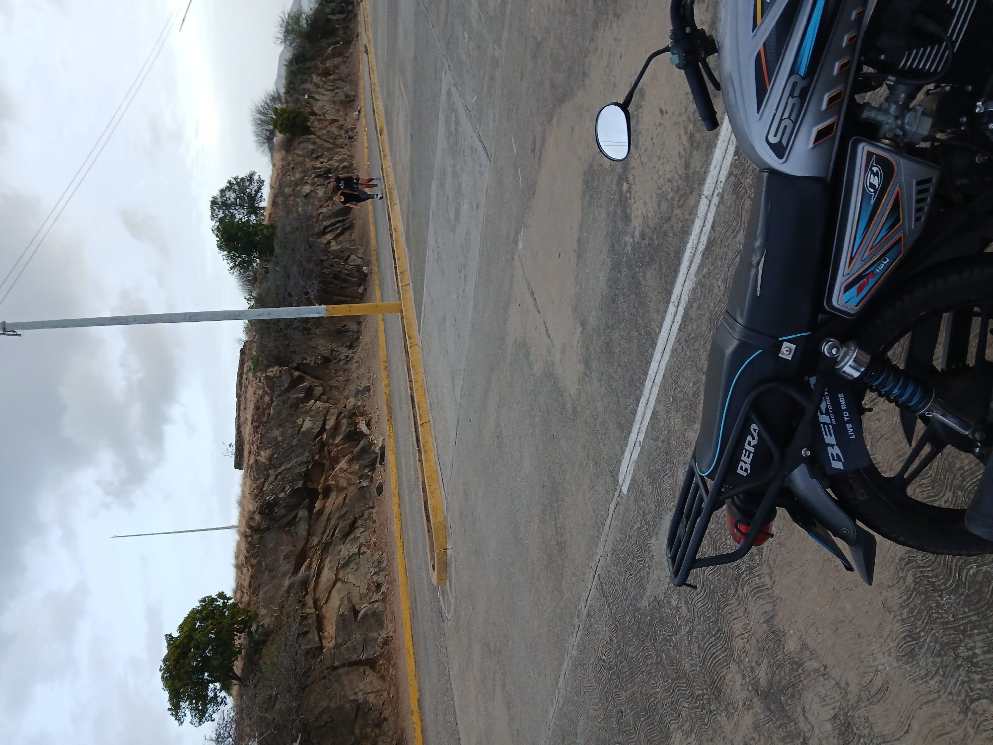 The image shows a motorcycle parked on a paved road with a yellow curb alongside a rocky cliff. In the background, there is a tall, thin pole. Two people are walking along the road. The motorcycle is partially in the foreground, with its right side visible. The sky is overcast. The location is Juan Griego, Venezuela.
