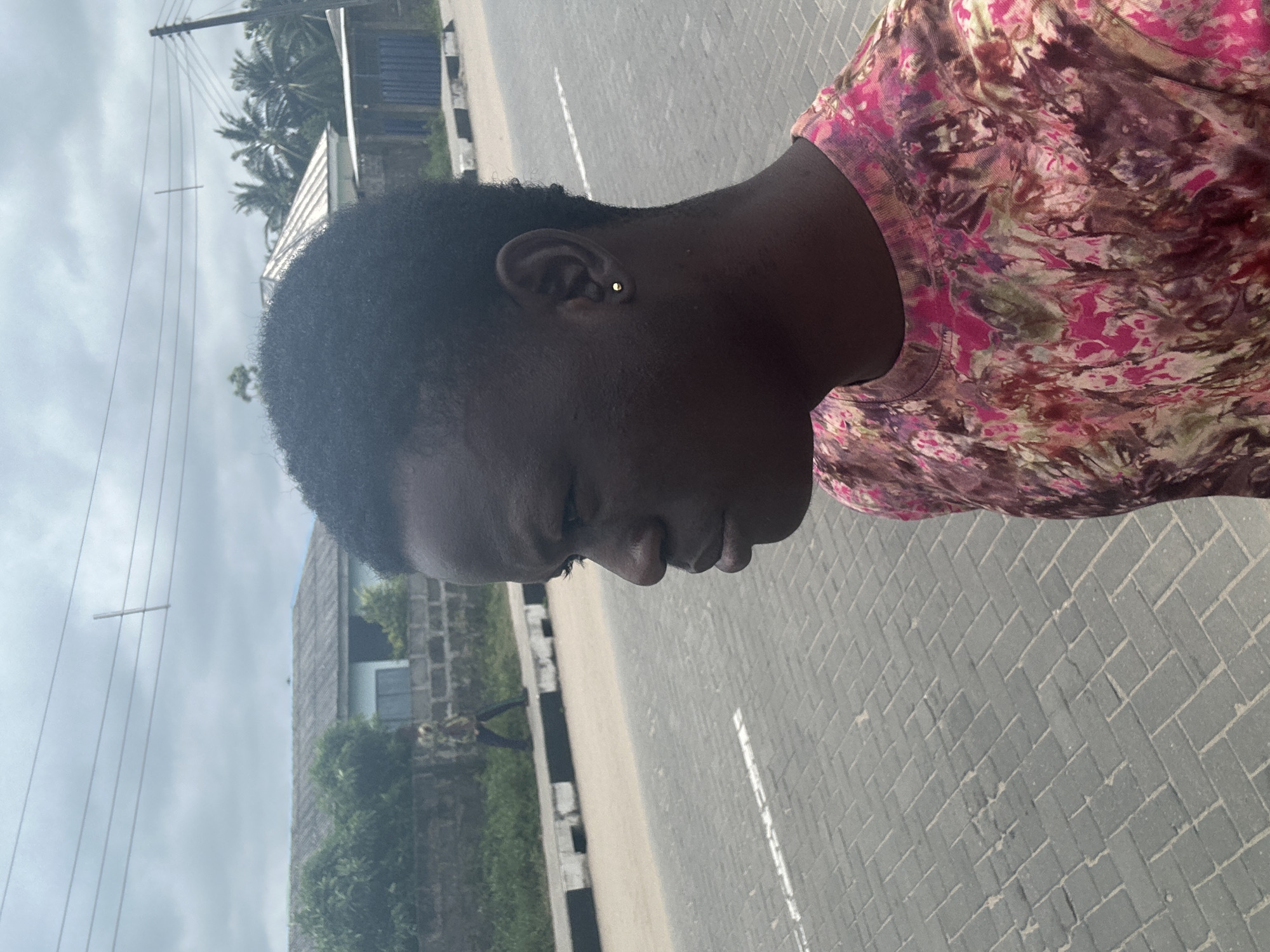 In Badagry, Nigeria, a young Black male is photographed from the chest up, facing to the left with his head slightly tilted down. He has short, dark, curly hair and is wearing a t-shirt with a multi-colored tie-dye pattern featuring shades of pink, purple, green, and brown. A small gold stud earring is visible in his left ear. The background shows a paved sidewalk with a white dashed line, a street with a raised curb, and in the distance, a low-rise building with a gray, corrugated metal roof, some trees, and power lines. A brick wall runs along the edge of the sidewalk, and a person in a colorful garment is walking away from the camera in the far distance. The sky is overcast with a cloudy, grayish-blue hue.