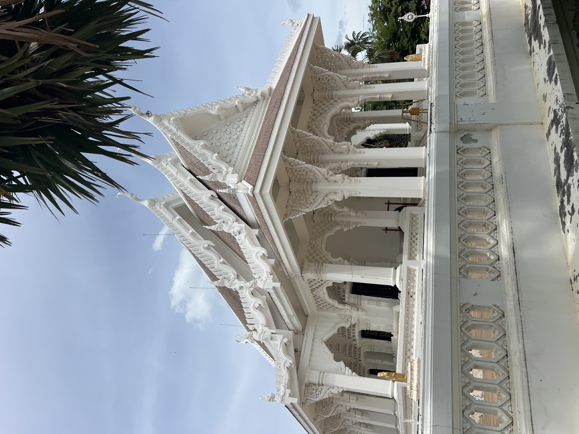 This is a vertical, low-angle, medium shot of a white ornate building, possibly a temple, in Baan Bo Phut, Thailand. The architecture features intricate white carvings on the eaves and pillars, with a brown tiled roof. The building has multiple levels and a colonnade. The sky is a clear light blue with a few wispy clouds. To the left of the frame, the fronds of a large palm tree hang down, obscuring some of the sky and building. The scene appears to be outdoors during the day, under fair weather conditions. The building shows some signs of wear and tear, with chipped paint and discoloration in places. The overall impression is one of serene architectural beauty in a tropical setting. No people or specific activities are visible. There is no visible text in the image.