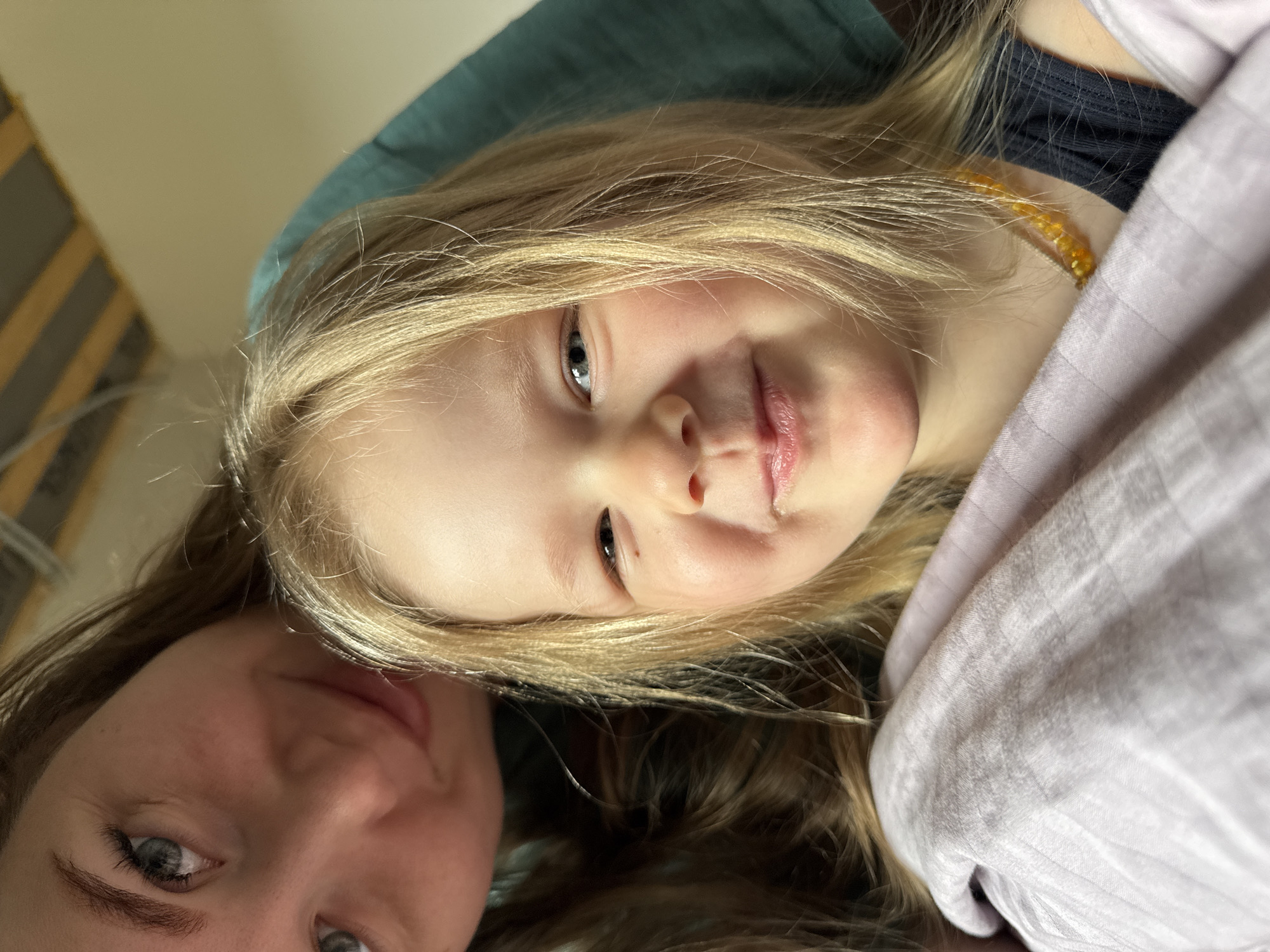 A close-up, indoor, selfie-style image shows two individuals, likely a mother and child, sharing a moment of rest or relaxation. The child, with fair blonde hair, is positioned prominently in the frame, looking directly at the camera with a slight smile. They appear to be wearing a dark-colored top and possibly a beaded necklace. Parts of a lavender-colored duvet or blanket can be observed around the child.
A woman with brown hair, possibly the child's mother, occupies the lower left corner of the frame. Her face is partially visible, also gazing towards the camera.
The background suggests an interior setting. A teal-colored cushion or headboard is visible, along with a light-colored wall in the top left corner. The image provides no information about the city of Ферма, Russia.